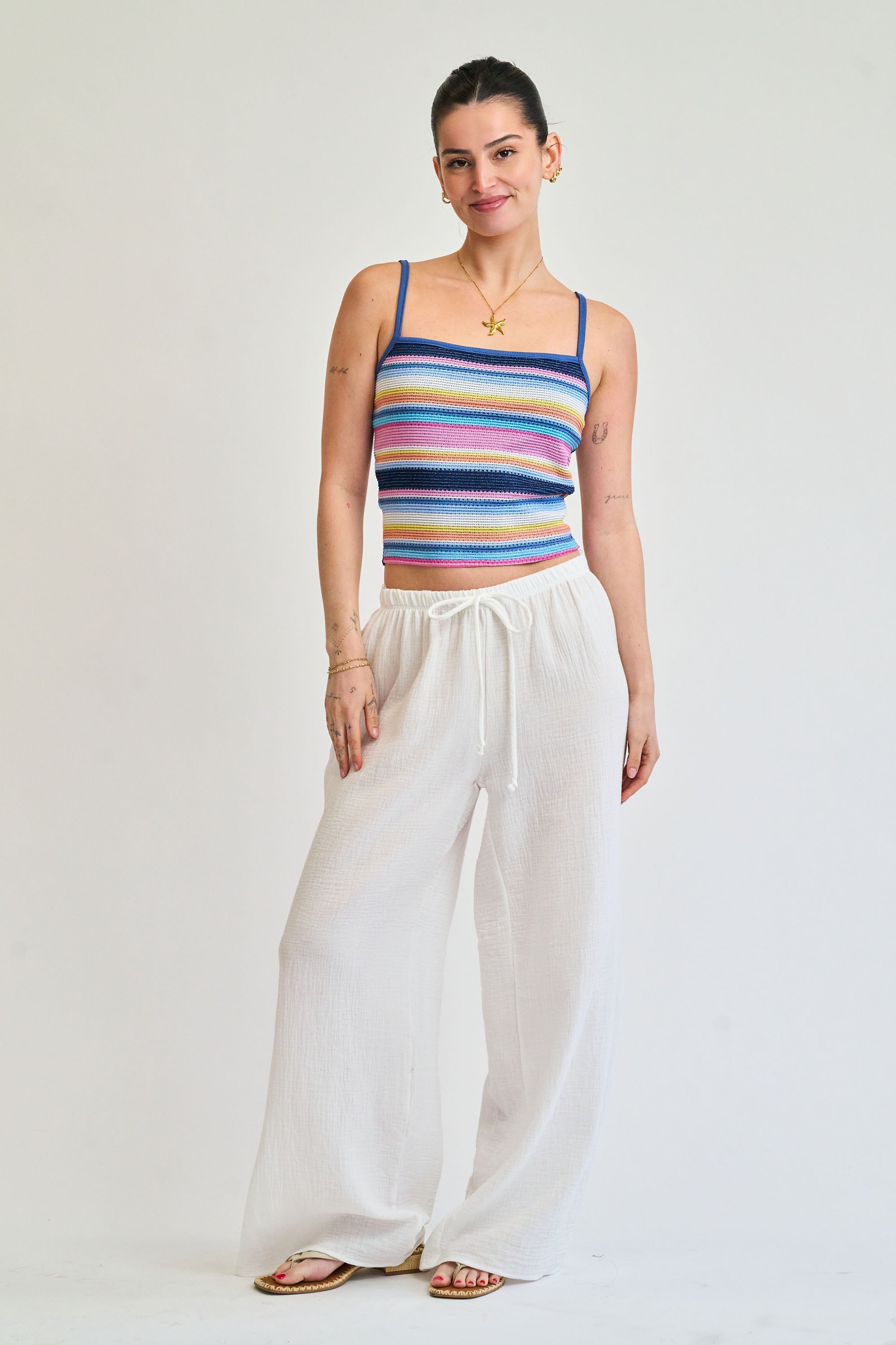 Woman wearing a colorful striped tank top and white pants on a plain background
