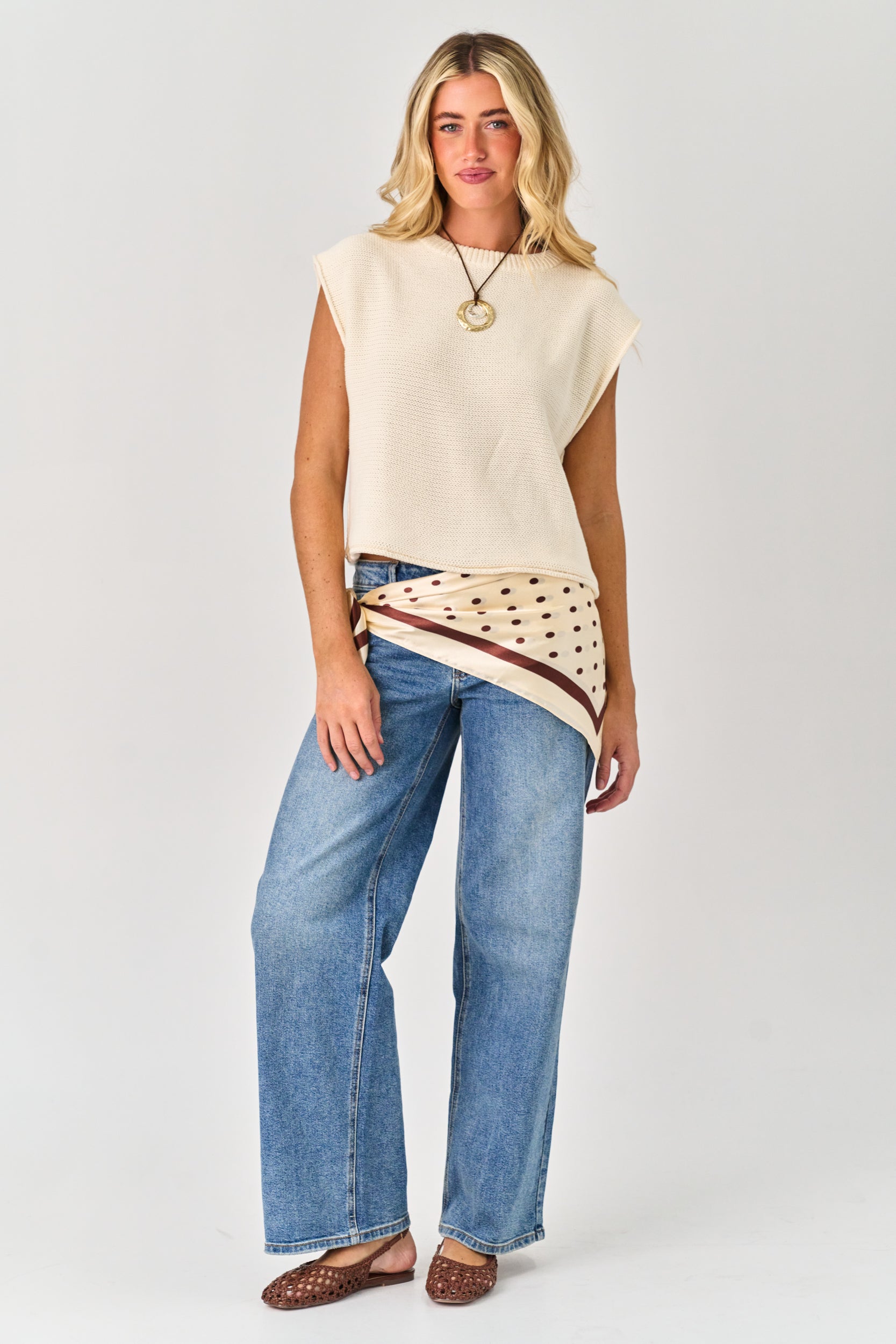 Woman wearing a cream sleeveless top with a patterned hem and blue jeans on a white background