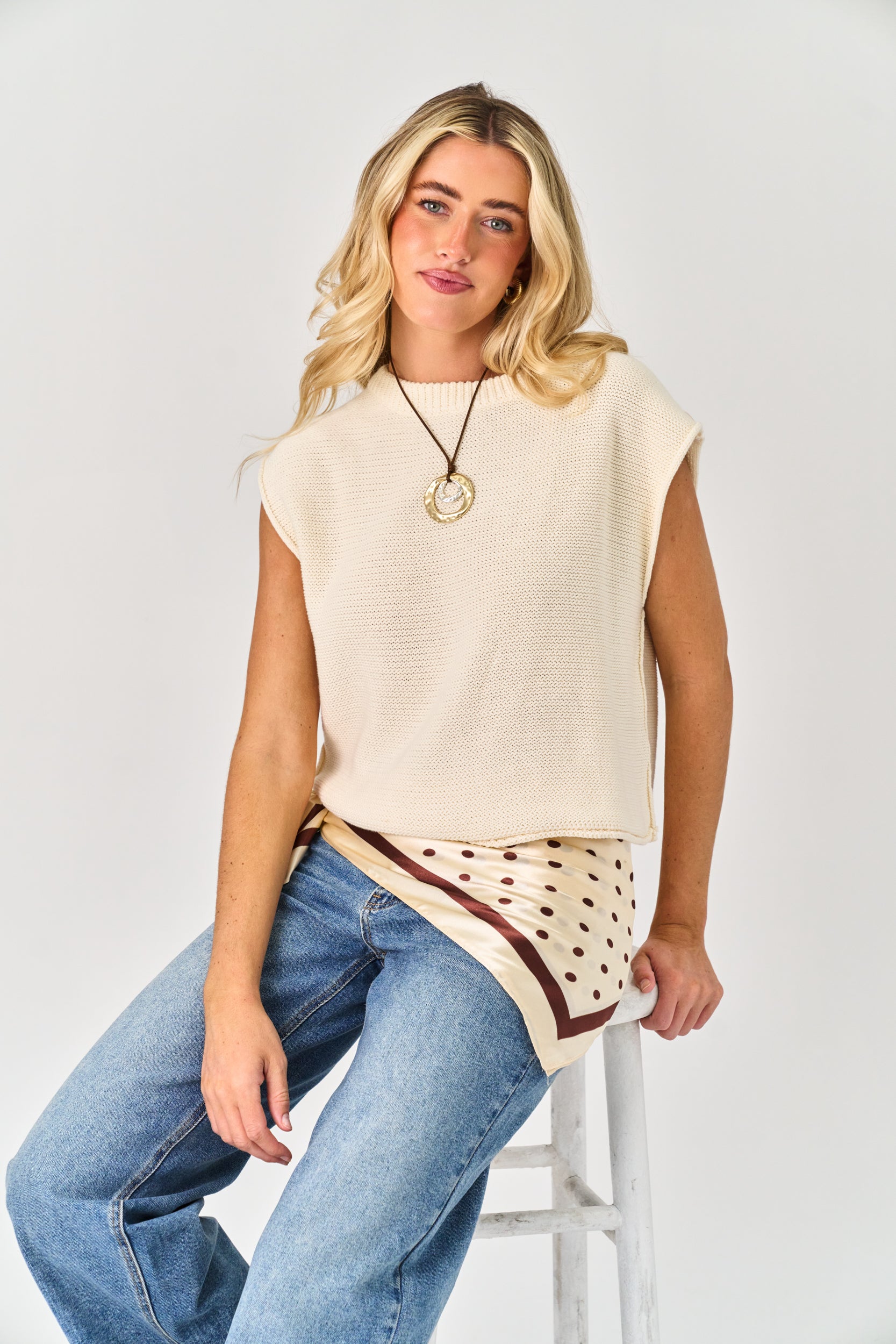 Woman wearing a beige sleeveless top with a patterned hem and blue jeans on a white background