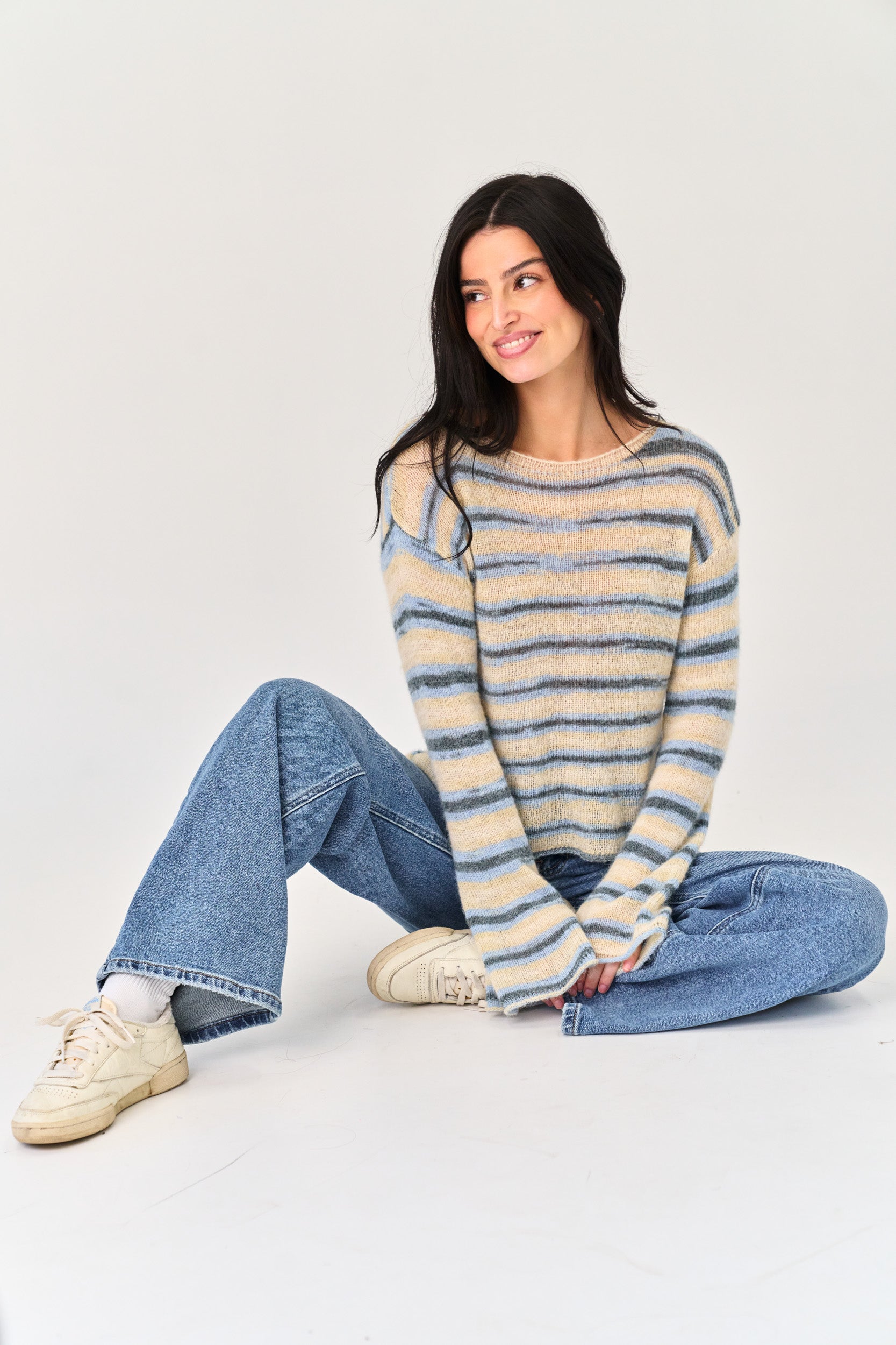 Woman wearing a striped sweater and jeans sitting on the floor against a plain background