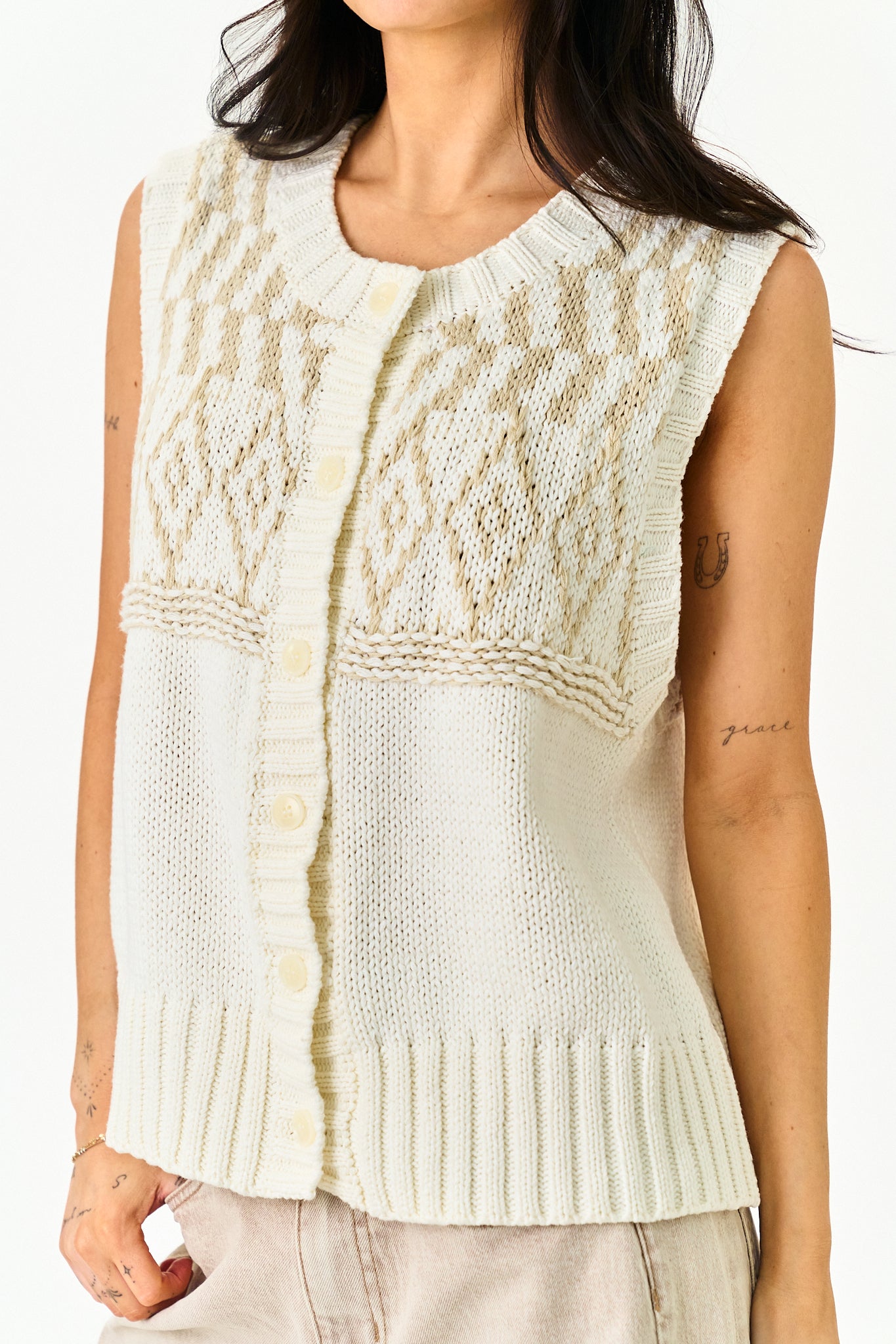 A woman wearing a sleeveless cream-colored knit vest with a buttoned front and textured cable-knit pattern, paired with light beige pants against a plain white background.