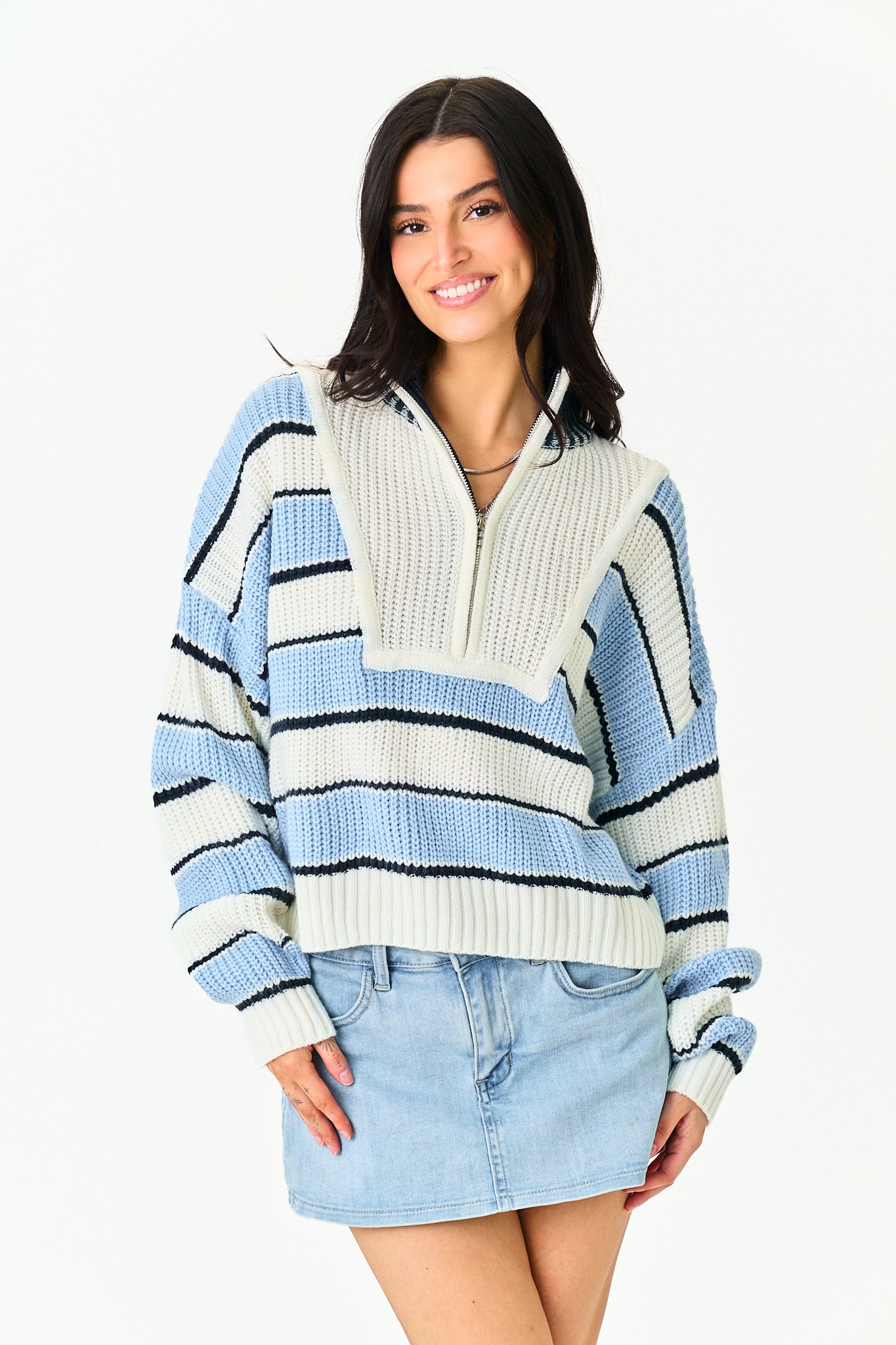 A woman wearing a light blue, white, and black striped ribbed knit half-zip sweater with a mock neck, paired with a light blue denim mini skirt, against a plain white background.
