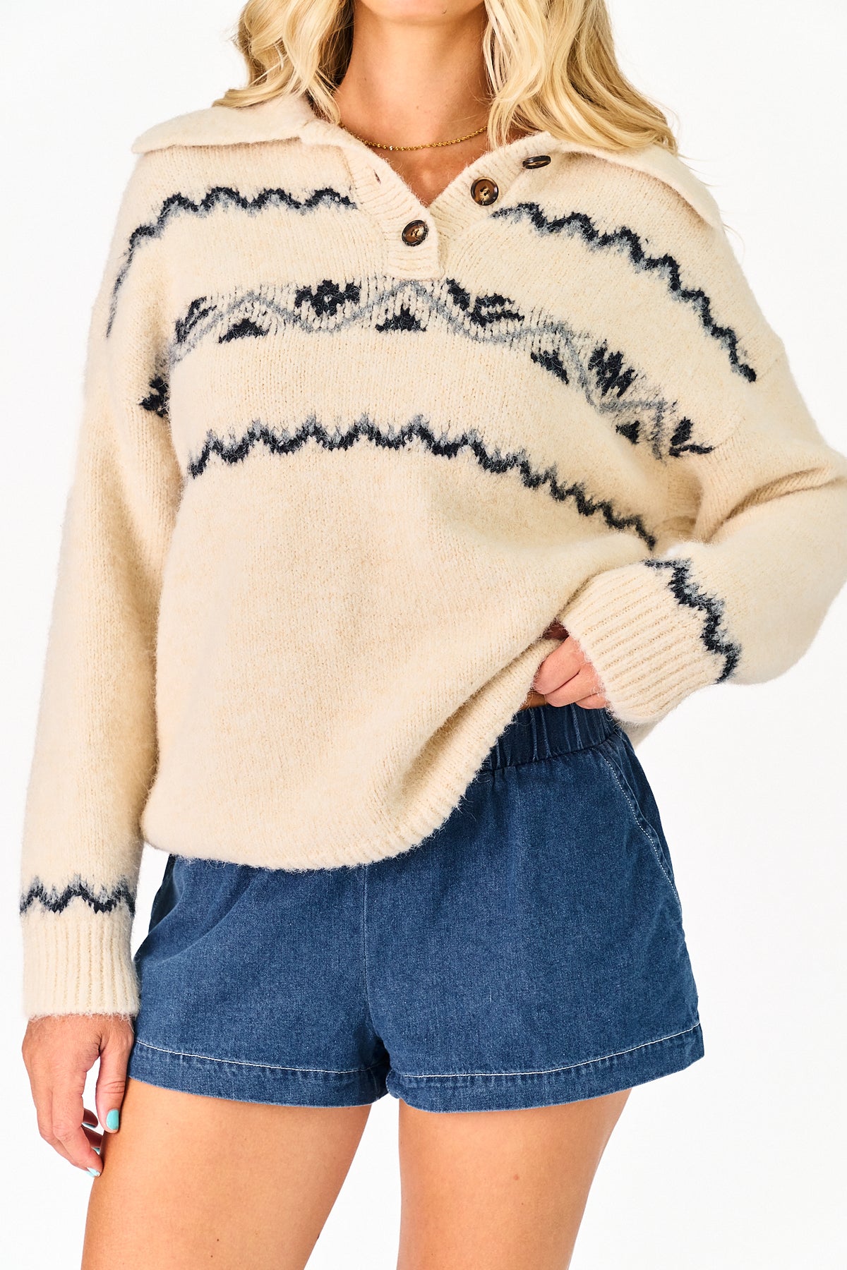 A woman wearing a fuzzy cream knit polo-style sweater with dark navy geometric and zigzag patterns across the chest and sleeves, paired with blue denim shorts against a plain white background.