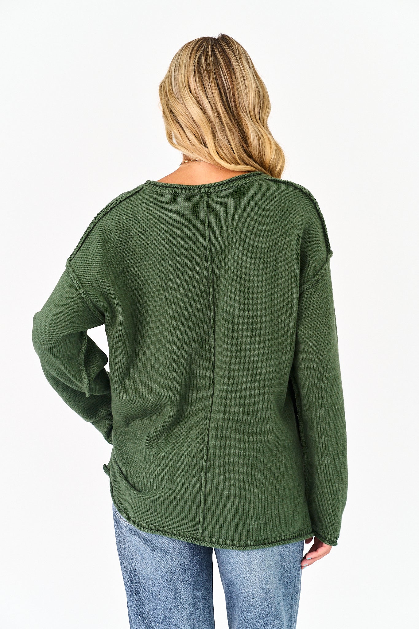 A woman viewed from the back wearing an olive green ribbed knit sweater and blue jeans against a plain white background.