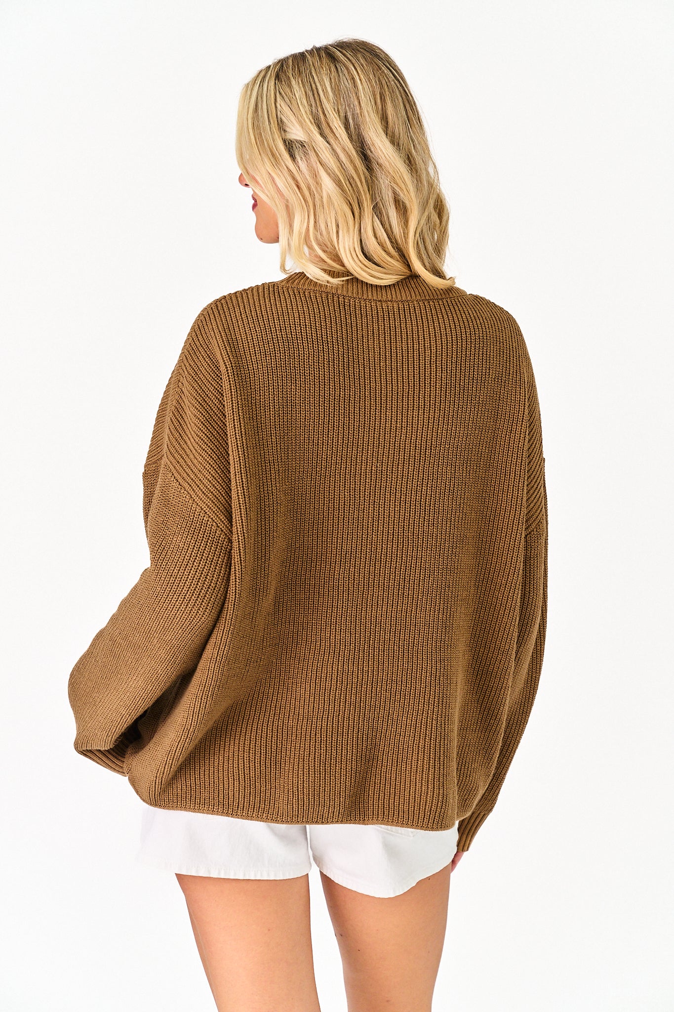 A woman shown from the back wearing an oversized ribbed knit sweater in a warm camel brown color, paired with white shorts, against a plain white background.