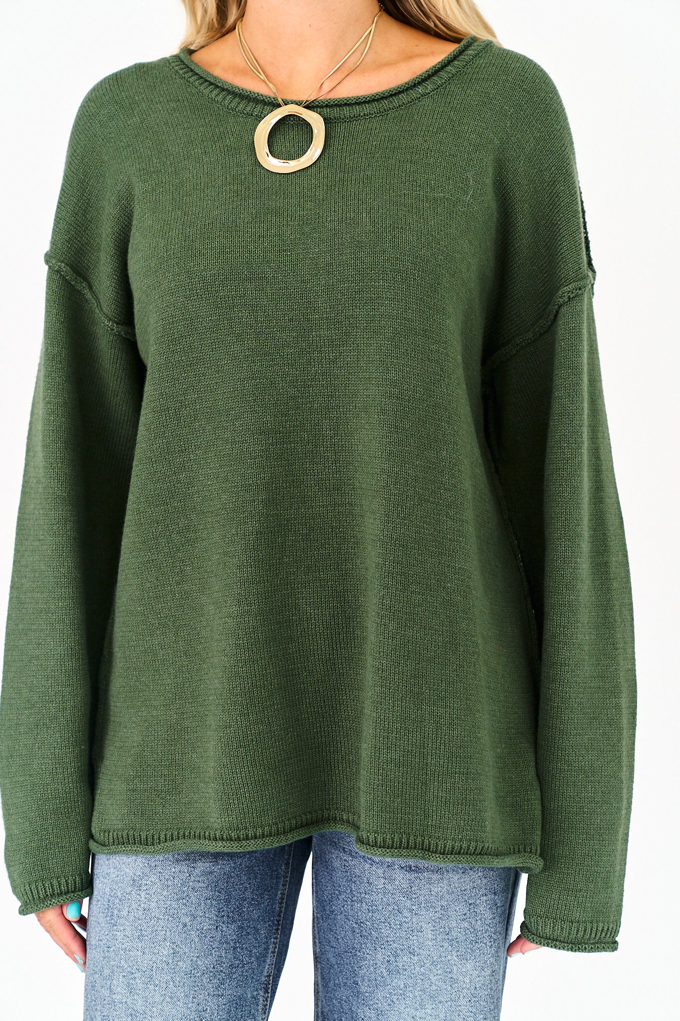 An olive green knit sweater with a wide crew neckline and a large gold circular pendant necklace worn by a model, paired with blue jeans.