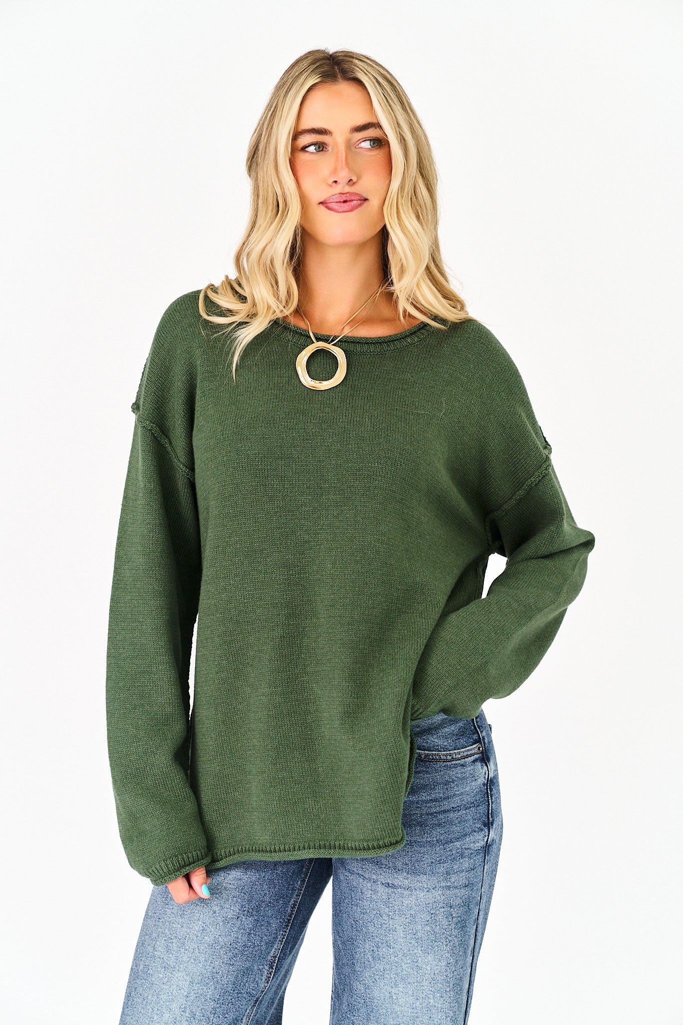A woman wearing an oversized olive green knit sweater with a wide crew neckline and a large gold circular pendant necklace, paired with blue jeans, against a plain white background.