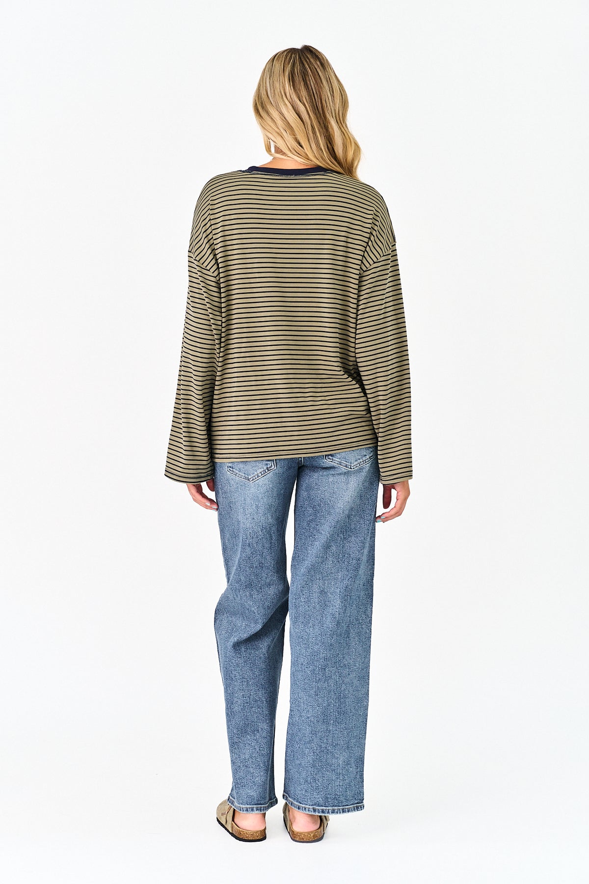 A woman viewed from the back wearing a long-sleeve olive green and beige striped top with blue jeans and sandals, against a plain white studio background.