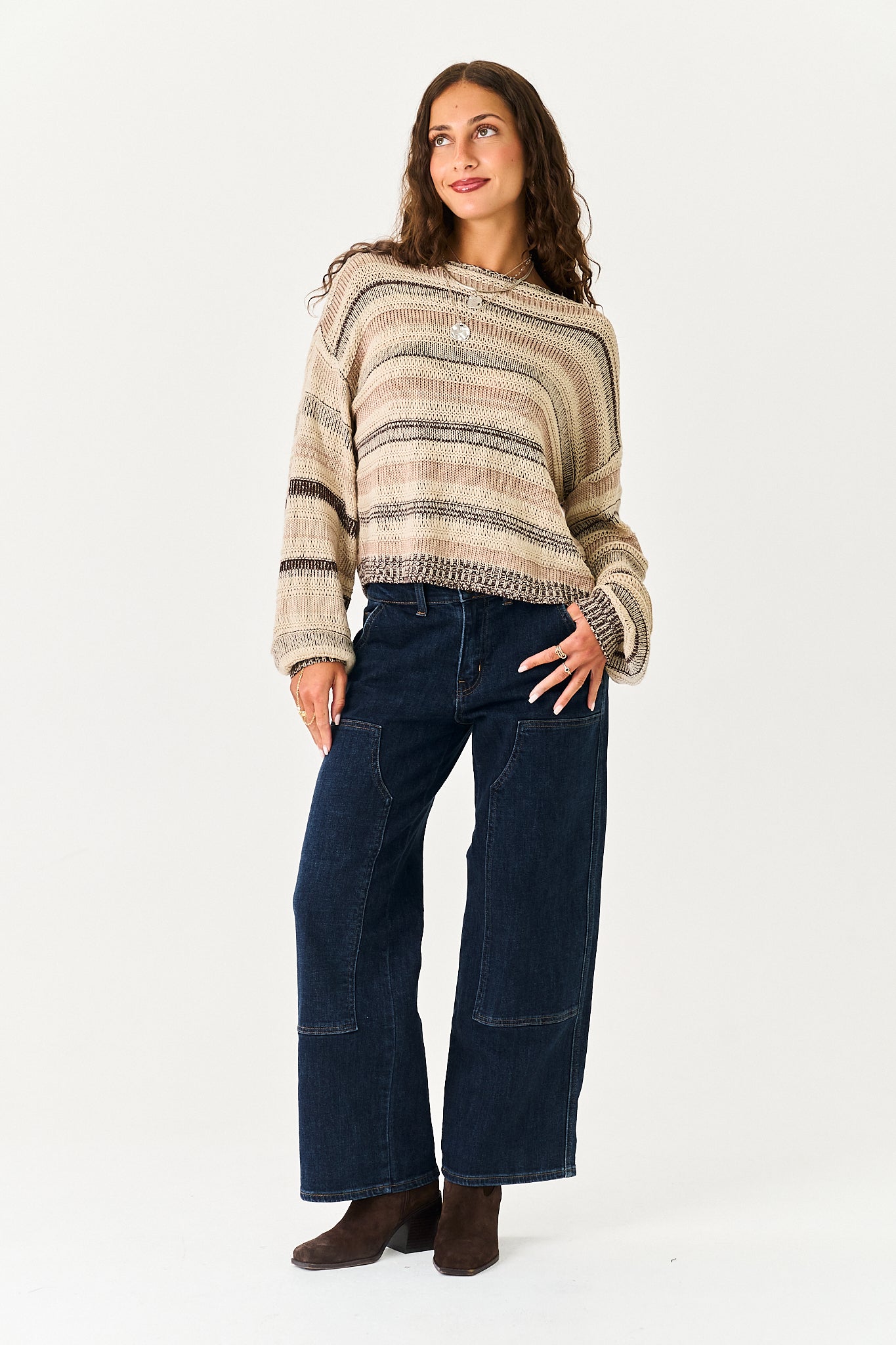 A woman modeling a beige and brown striped knit sweater with wide-leg dark blue jeans and brown boots, standing against a plain white studio background.