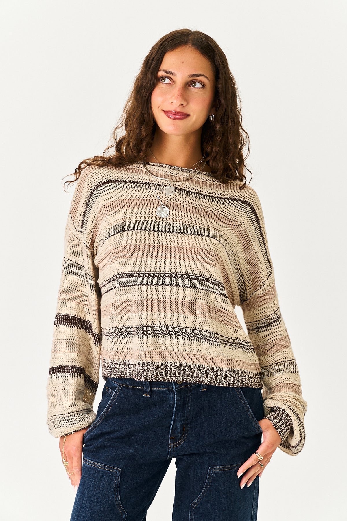 A woman wearing a beige and brown striped knit sweater with a crew neck and buttoned cuffs, paired with dark blue jeans, against a plain light background.