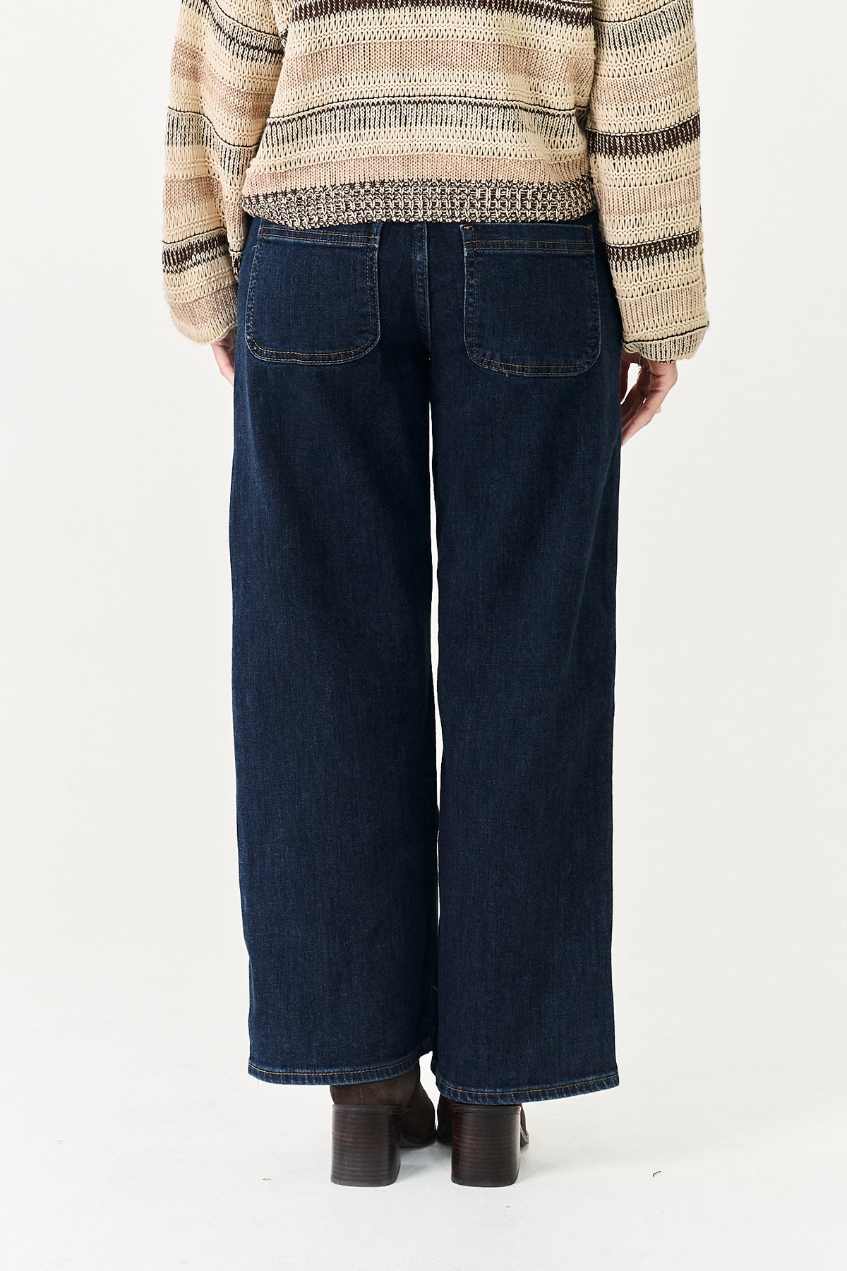Back view of a person wearing dark blue wide-leg denim jeans with back patch pockets, paired with a beige and brown striped knit sweater and dark brown boots, against a plain white studio background.