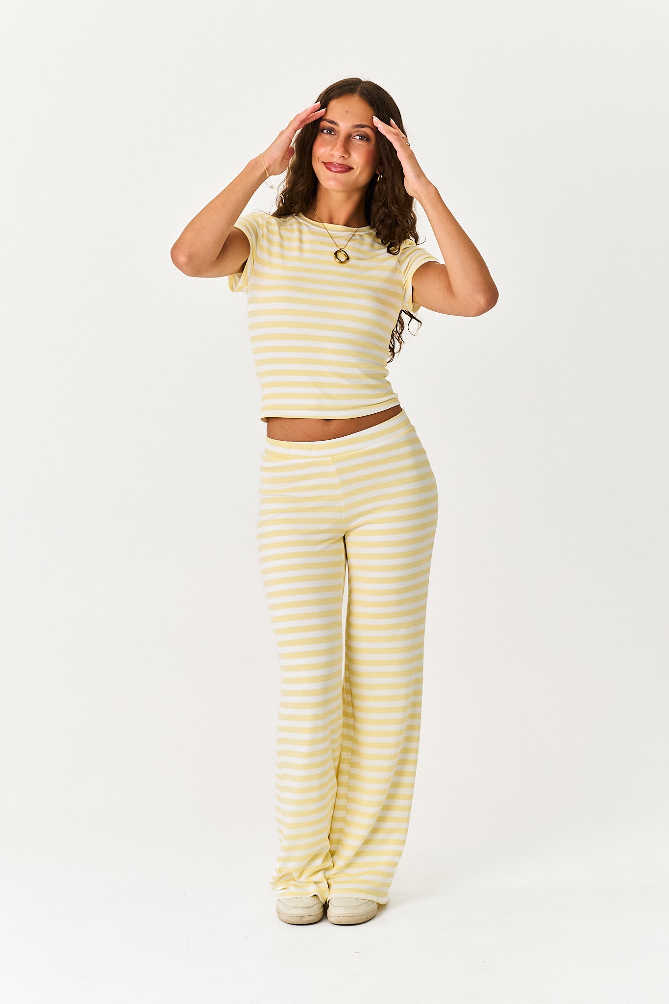 A woman modeling a matching two-piece loungewear set consisting of a short-sleeve crop top and wide-leg pants, both in a pale yellow and white horizontal stripe pattern, against a plain white studio background.