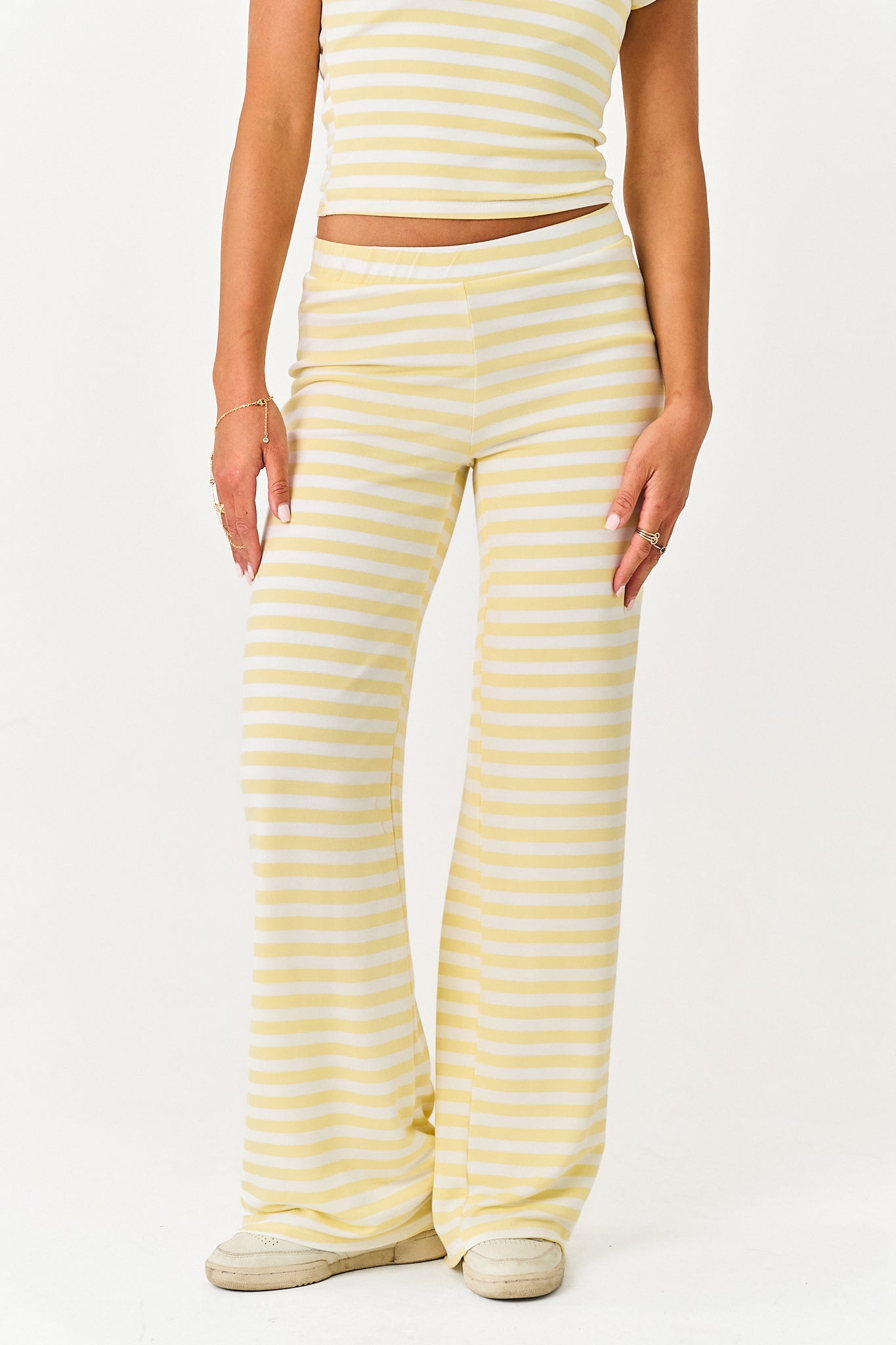 A model wearing pale yellow and white horizontally striped wide-leg pants, paired with a matching striped crop top and beige sneakers, against a plain white studio background.
