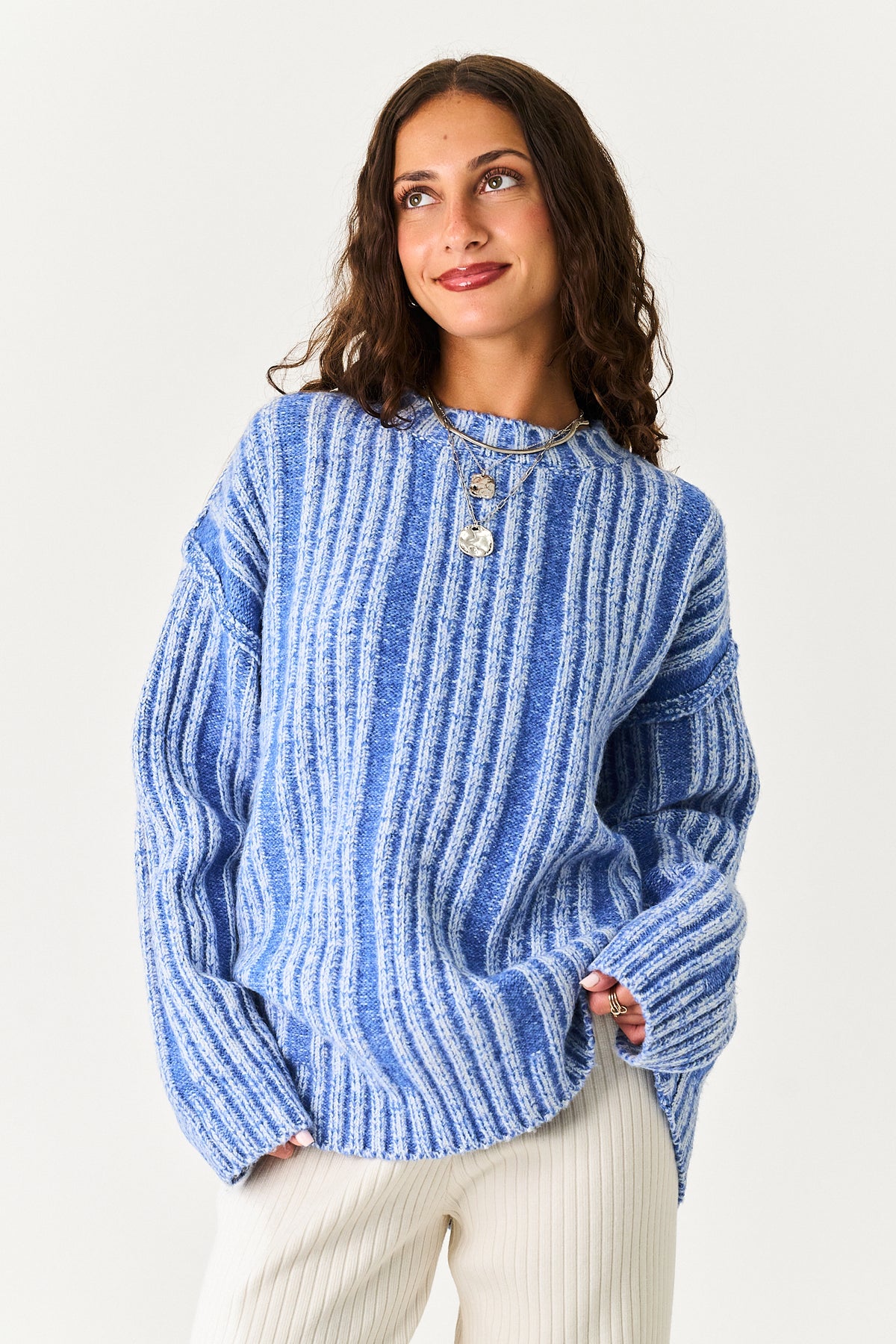 A woman wearing a blue and white vertically striped knit sweater with a buttoned neckline, paired with cream-colored pants, against a plain light background.