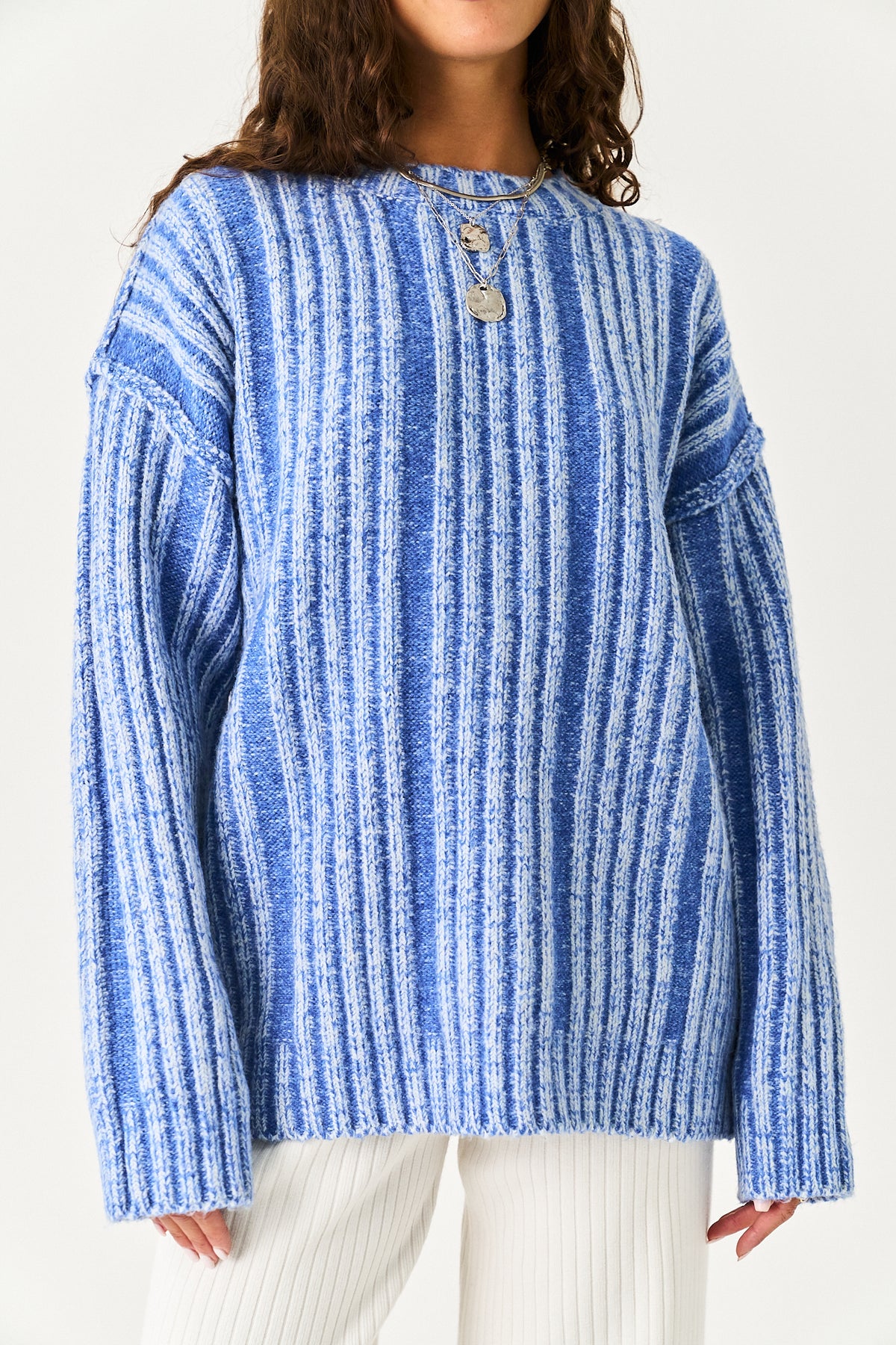 A person wearing a blue and white vertically striped knit sweater with a crew neck and decorative buttons, paired with white pants, against a plain light background.