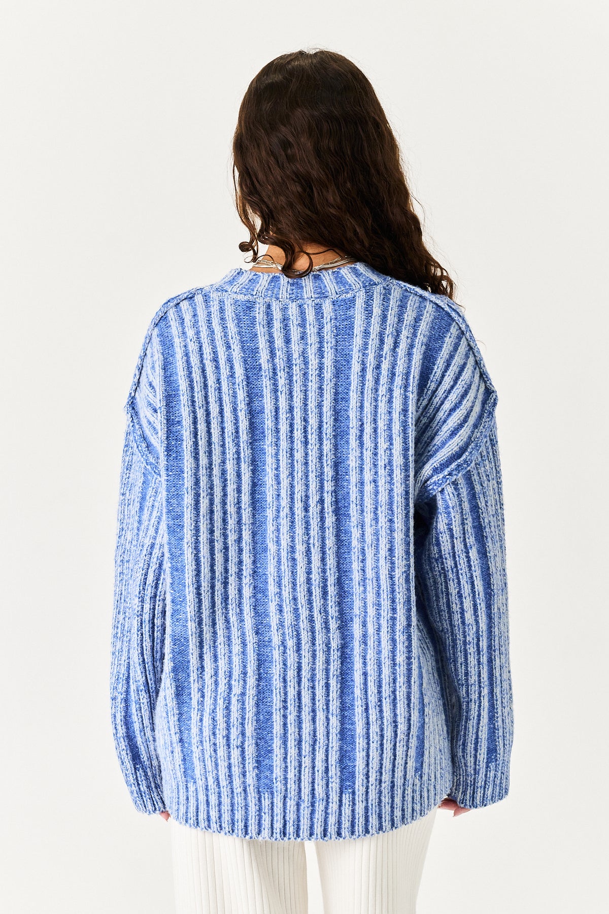 A woman viewed from behind wearing a blue and white vertically striped knit sweater and white pants against a plain off-white background.