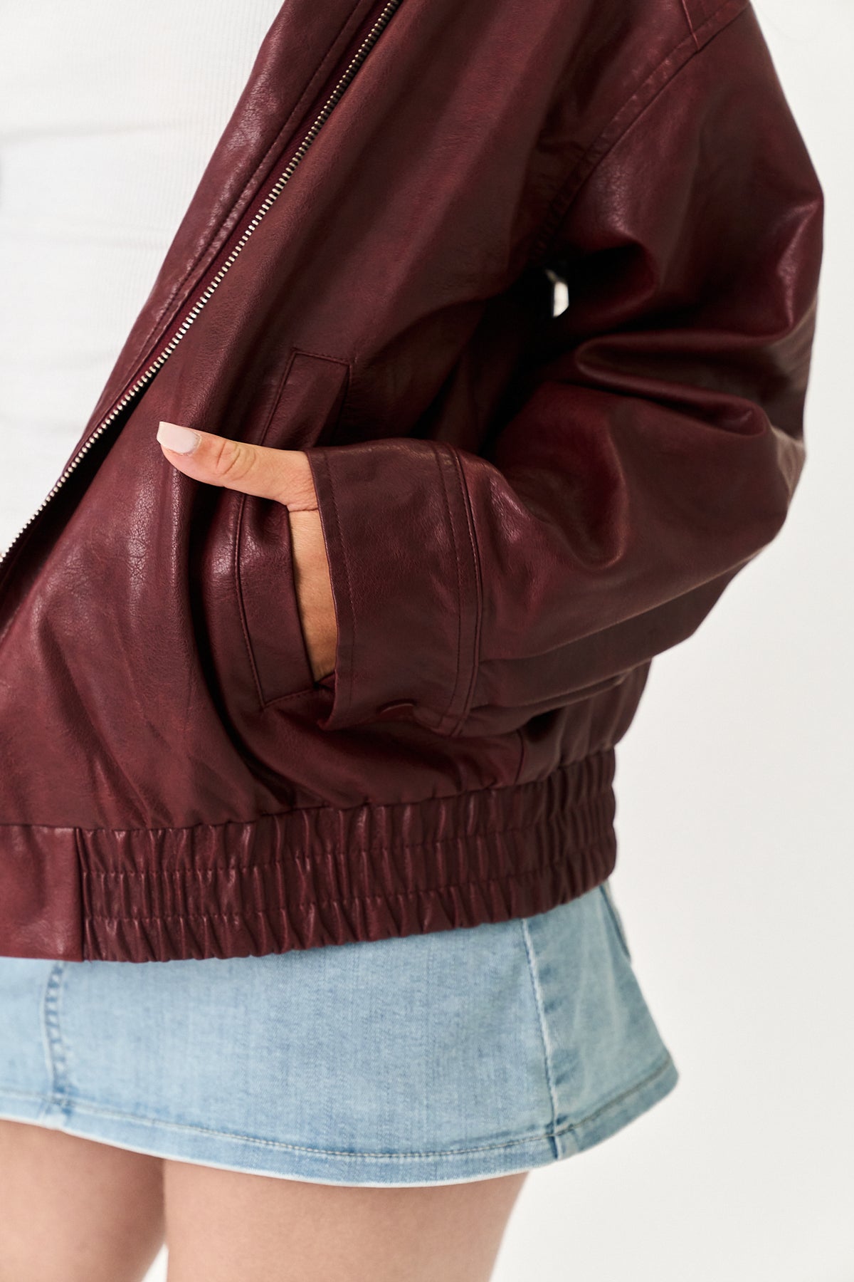 A close-up of a person wearing a dark burgundy faux leather jacket with a zippered chest and elasticated hem, paired with a light blue denim skirt.