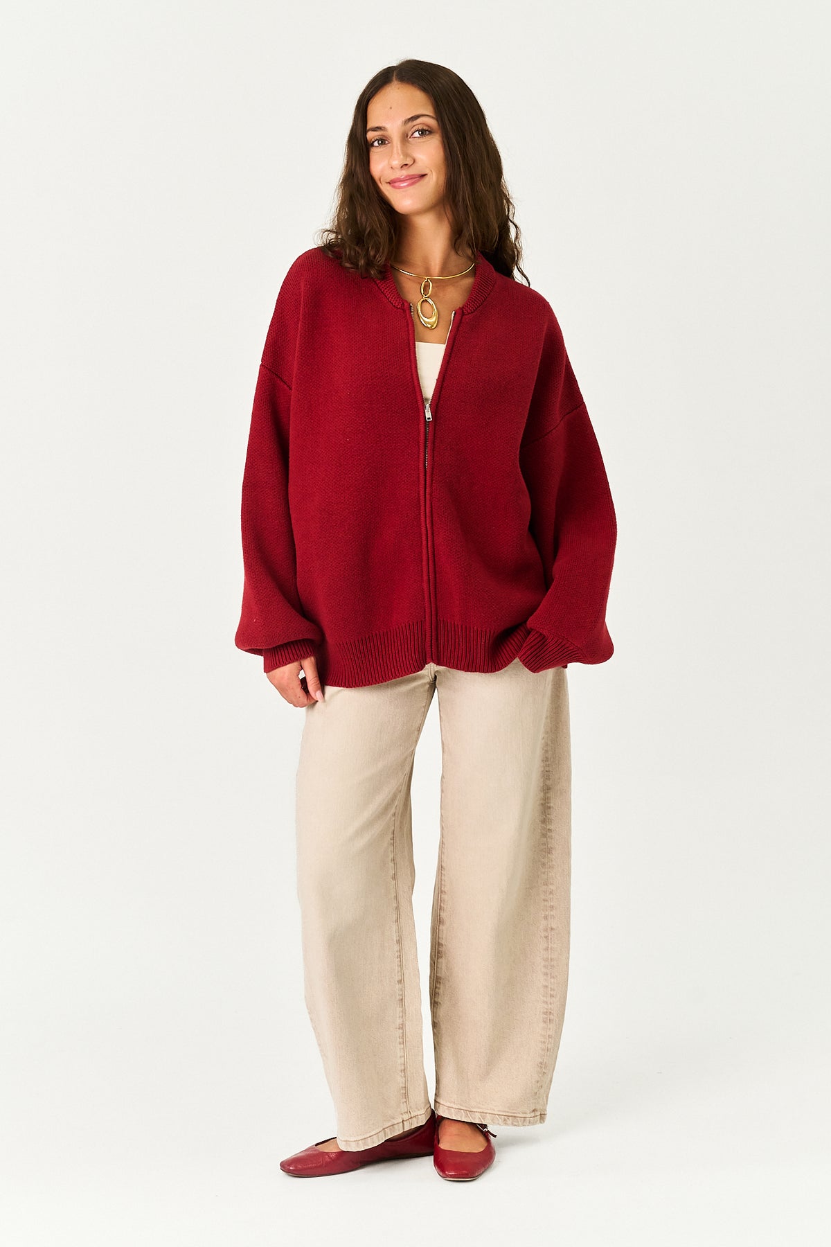 A woman modeling an oversized deep red zip-up cardigan with wide beige pants and red flats, standing against a plain light gray studio background.