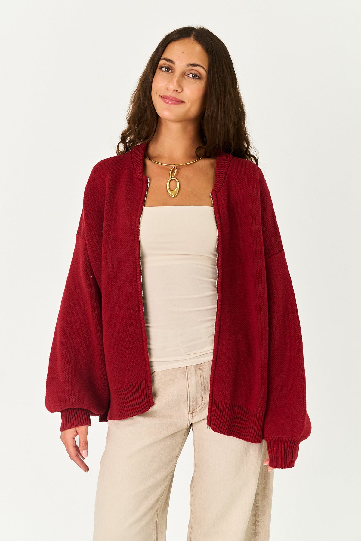 A woman modeling an oversized, deep burgundy knit cardigan over a cream strapless top and light beige pants, set against a plain light gray studio background.