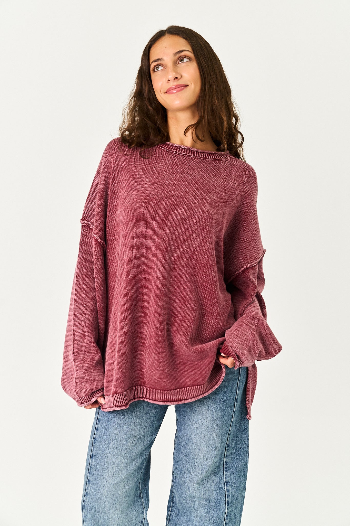A woman wearing a washed, oversized dusty rose knit sweater and blue jeans, standing against a plain white studio background.