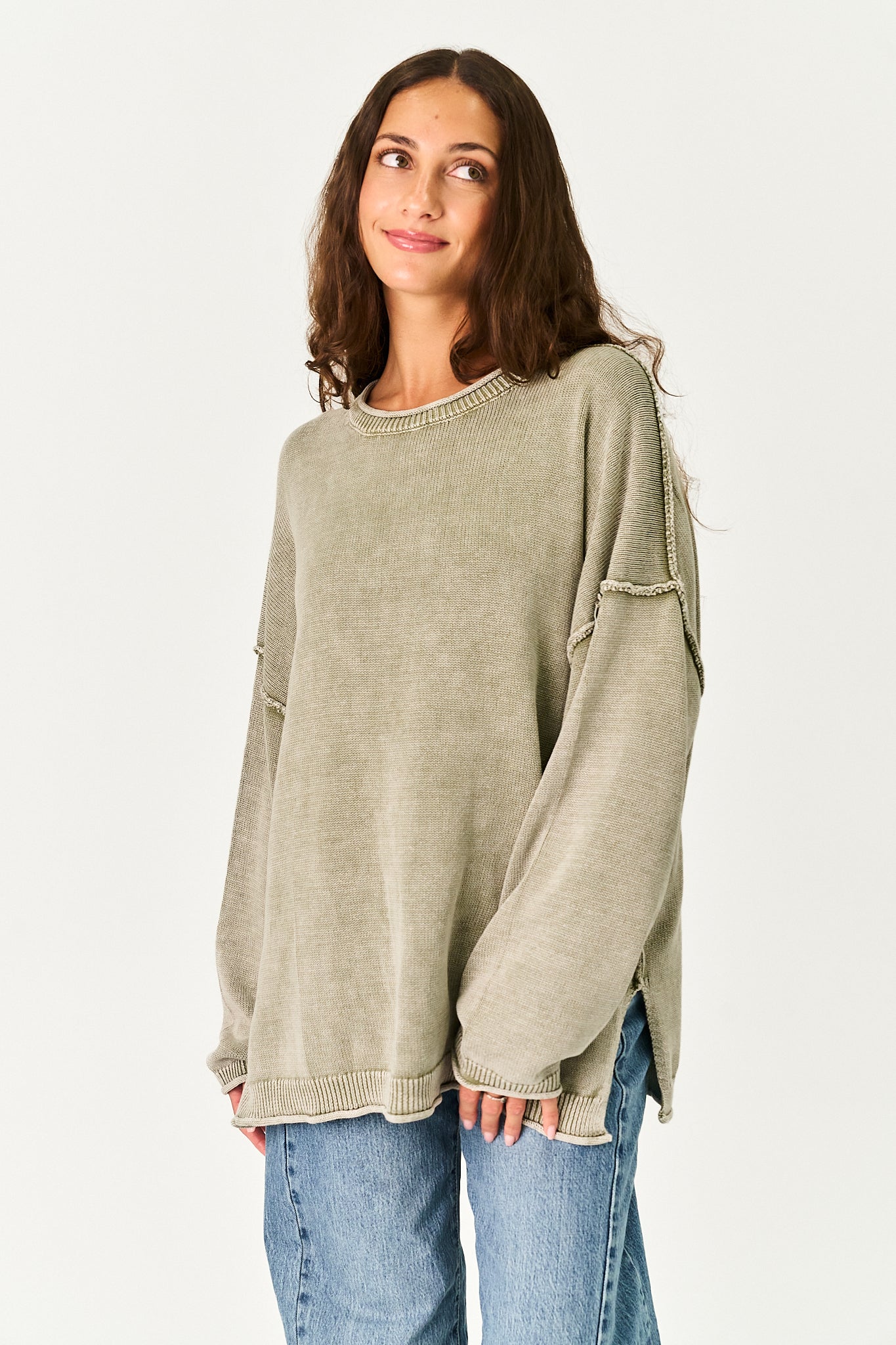 A woman wearing an oversized, long-sleeve knit sweater in a light sage green color, paired with blue jeans, against a plain white studio background.