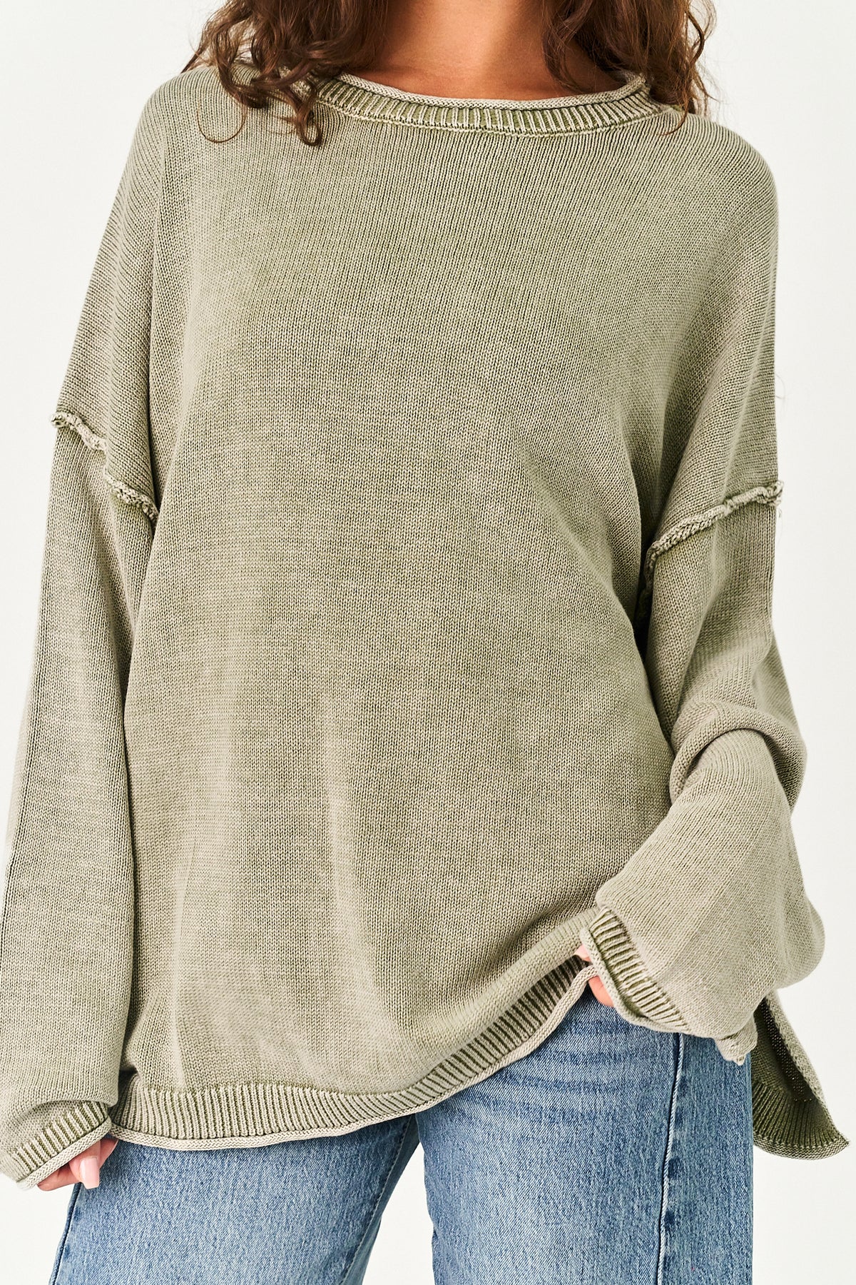 A person wearing an oversized ribbed knit sweater in a muted sage green color, paired with blue jeans, shown from the shoulders down against a plain light background.