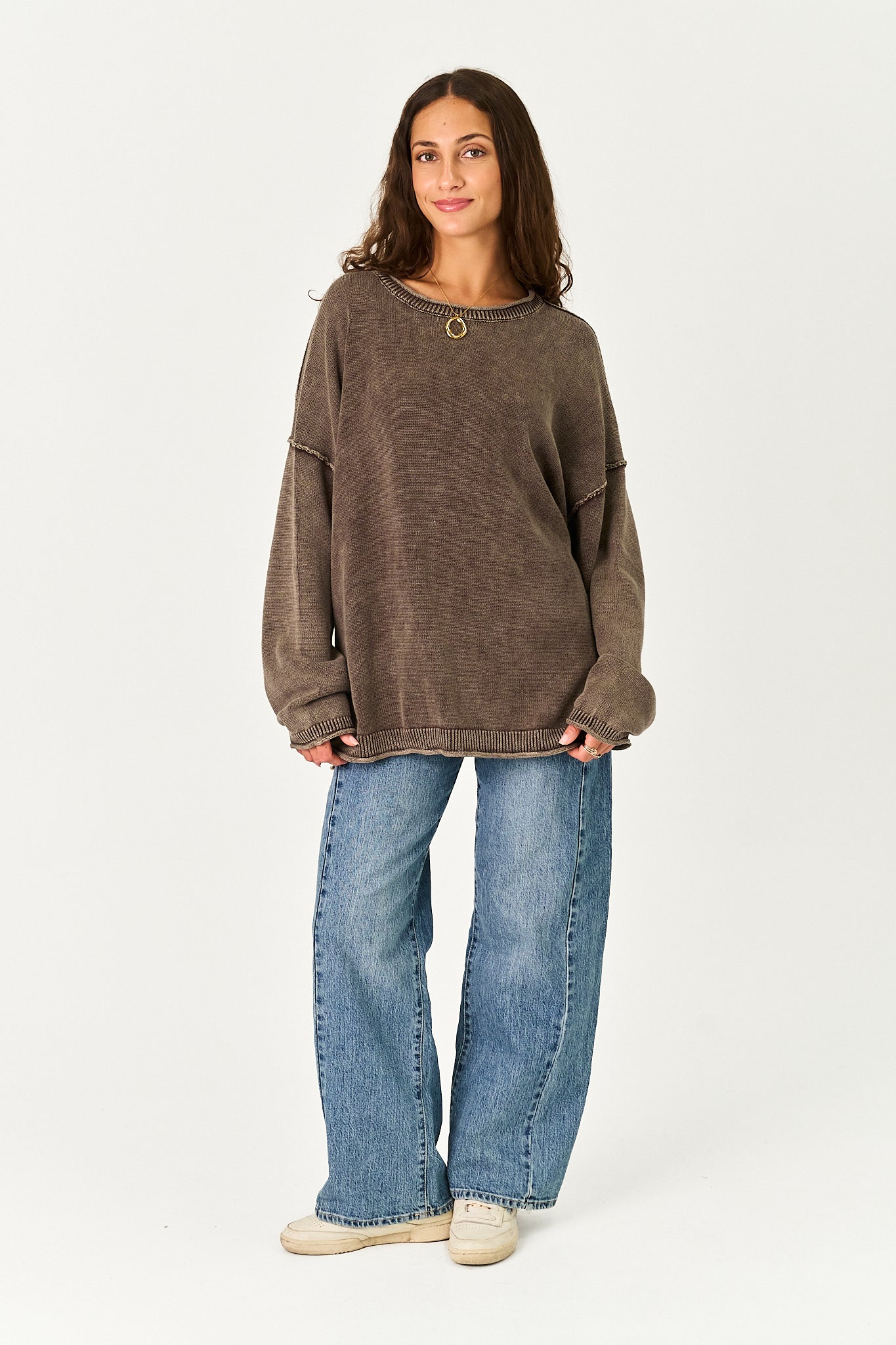 A woman modeling a brown washed, oversized crewneck sweatshirt with a single gold button closure, paired with wide-leg blue jeans and beige sneakers, against a plain white studio background.