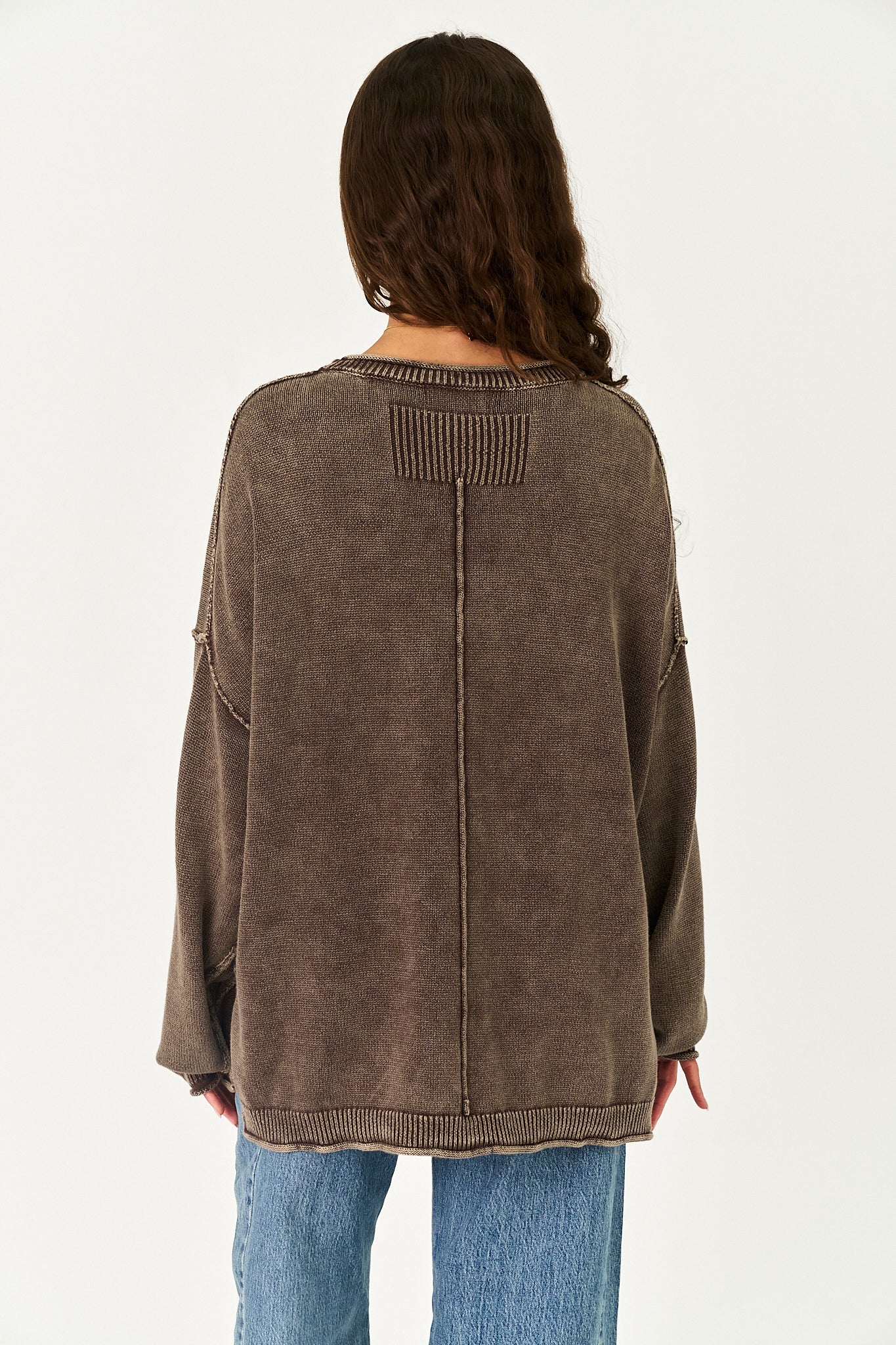 A woman viewed from the back wearing a brown ribbed knit sweater and blue jeans against a plain white background.