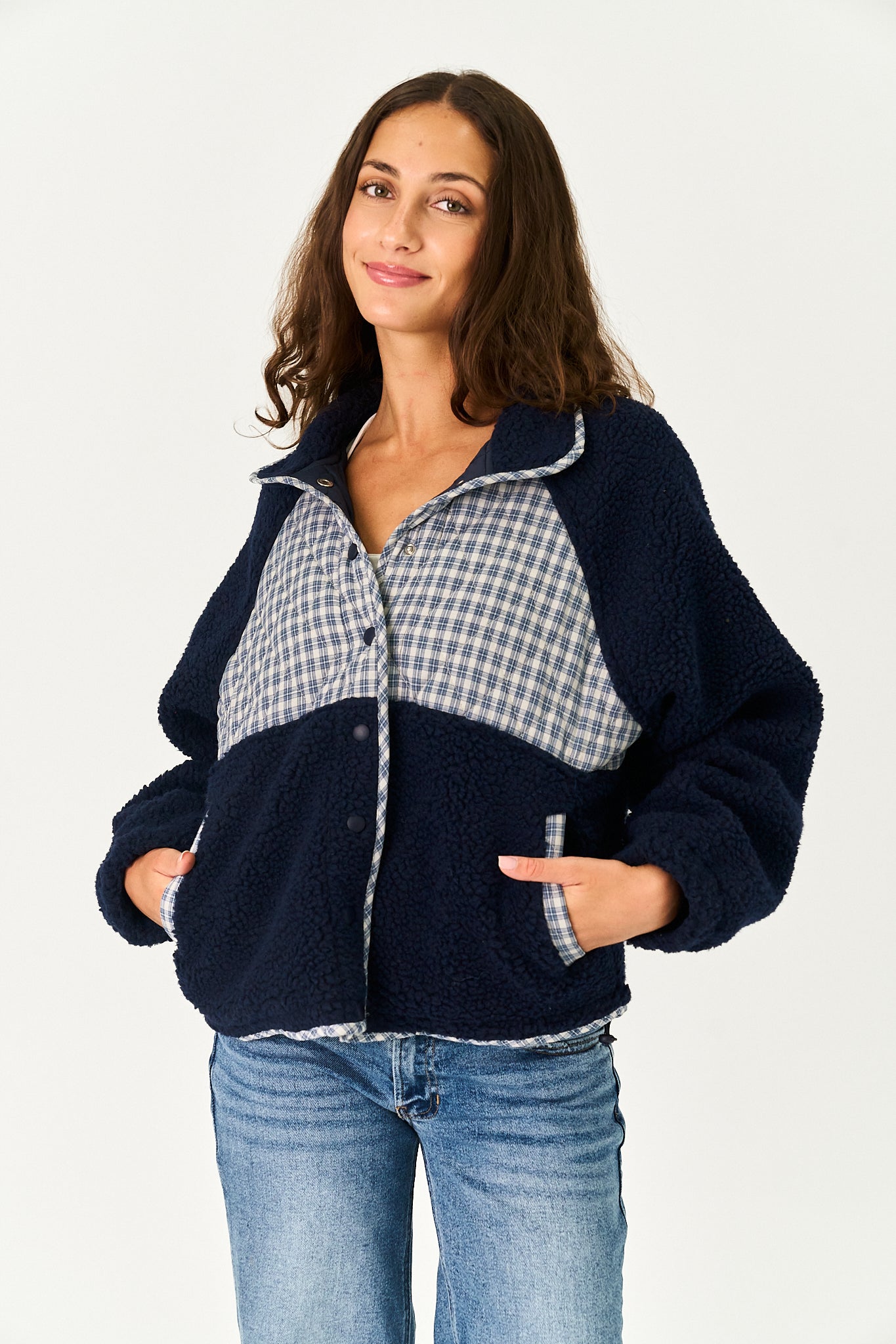 A woman modeling a navy blue sherpa fleece jacket with a light blue and white gingham plaid yoke panel and matching plaid trim, paired with blue jeans against a plain white background.