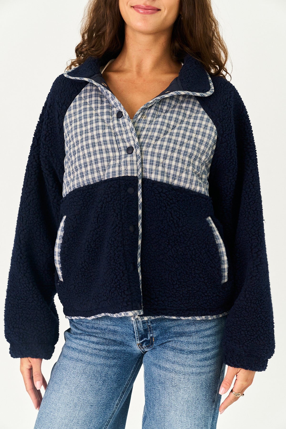 A woman wearing a navy blue sherpa fleece jacket with a light blue and white gingham plaid yoke panel and matching plaid trim, paired with blue jeans against a plain white background.