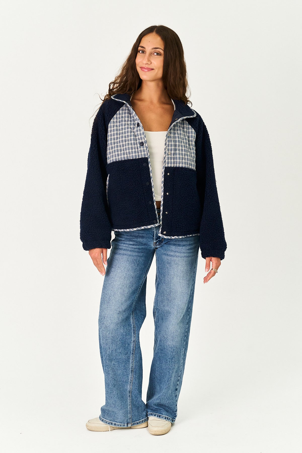 A woman modeling a navy blue fleece jacket with light blue and white gingham patterned yoke panels, paired with blue jeans and white sneakers, against a plain white studio background.