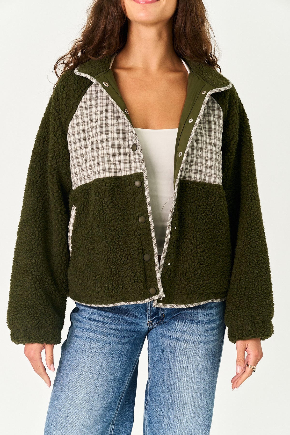 A woman wearing an olive green sherpa fleece jacket with beige and white gingham plaid panels on the chest, styled with a white tank top and blue jeans against a plain white background.