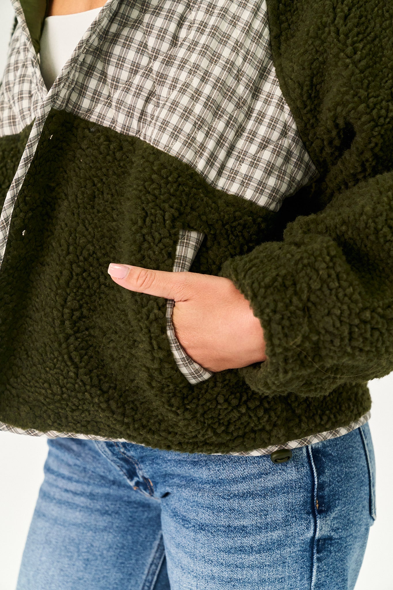 Close-up of a person wearing a color-block fleece jacket with an olive green sherpa body, a black and white plaid collar and shoulder panel, and blue jeans.