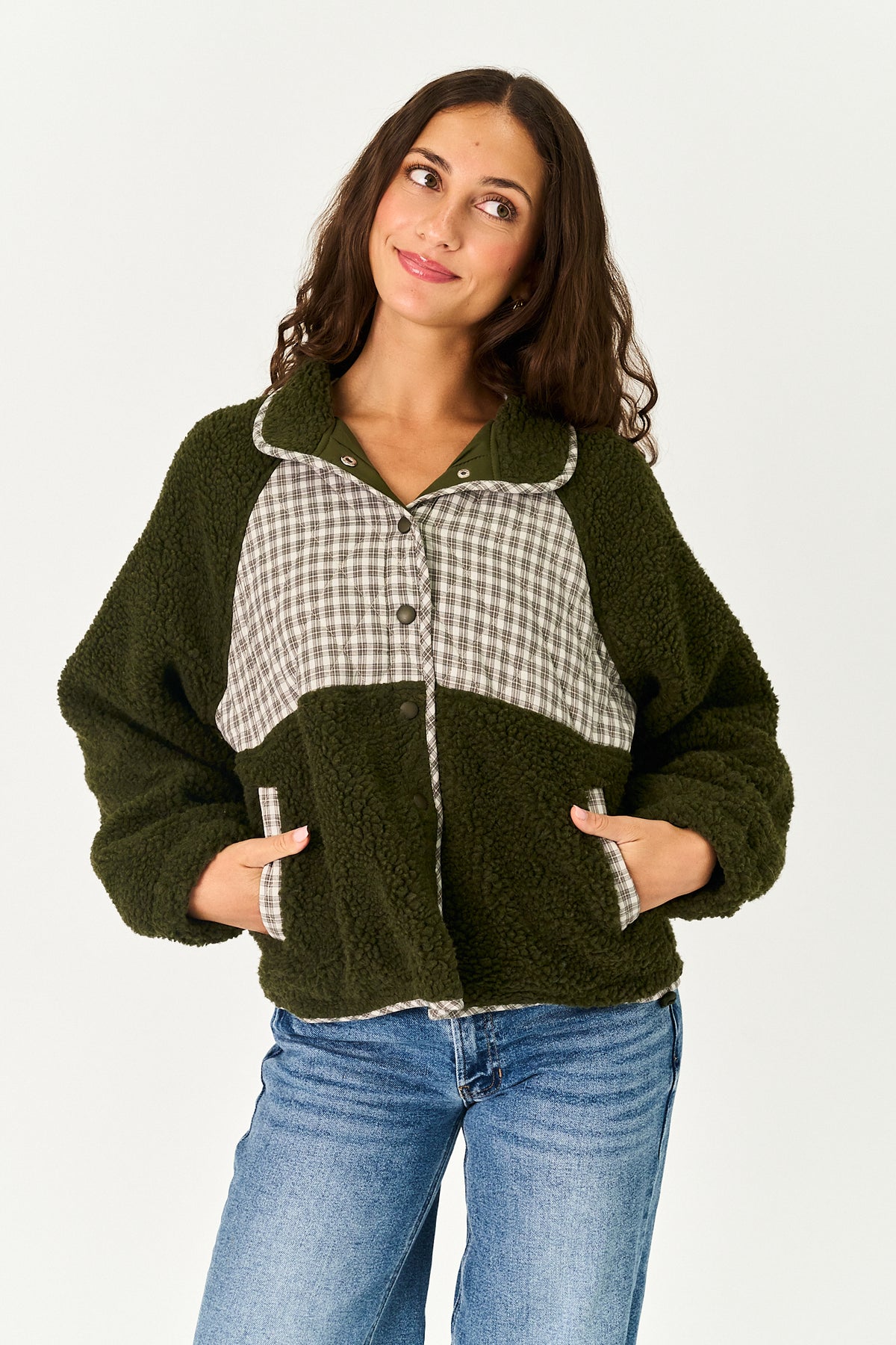 A woman wearing a dark olive green sherpa fleece jacket with a beige and white gingham check panel on the chest, paired with blue jeans, against a plain white studio background.