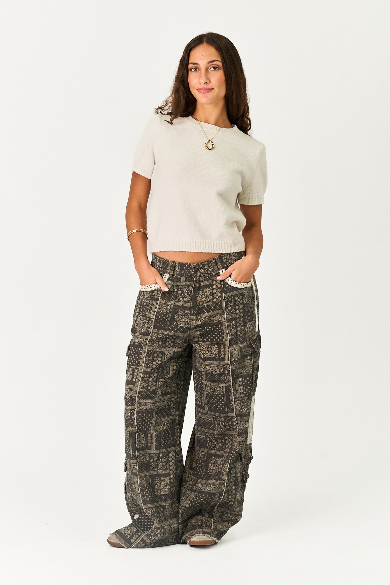 A woman modeling a pair of wide-leg cargo pants with a dark gray and black geometric patchwork pattern, paired with a cream short-sleeve crop top, against a plain white studio background.