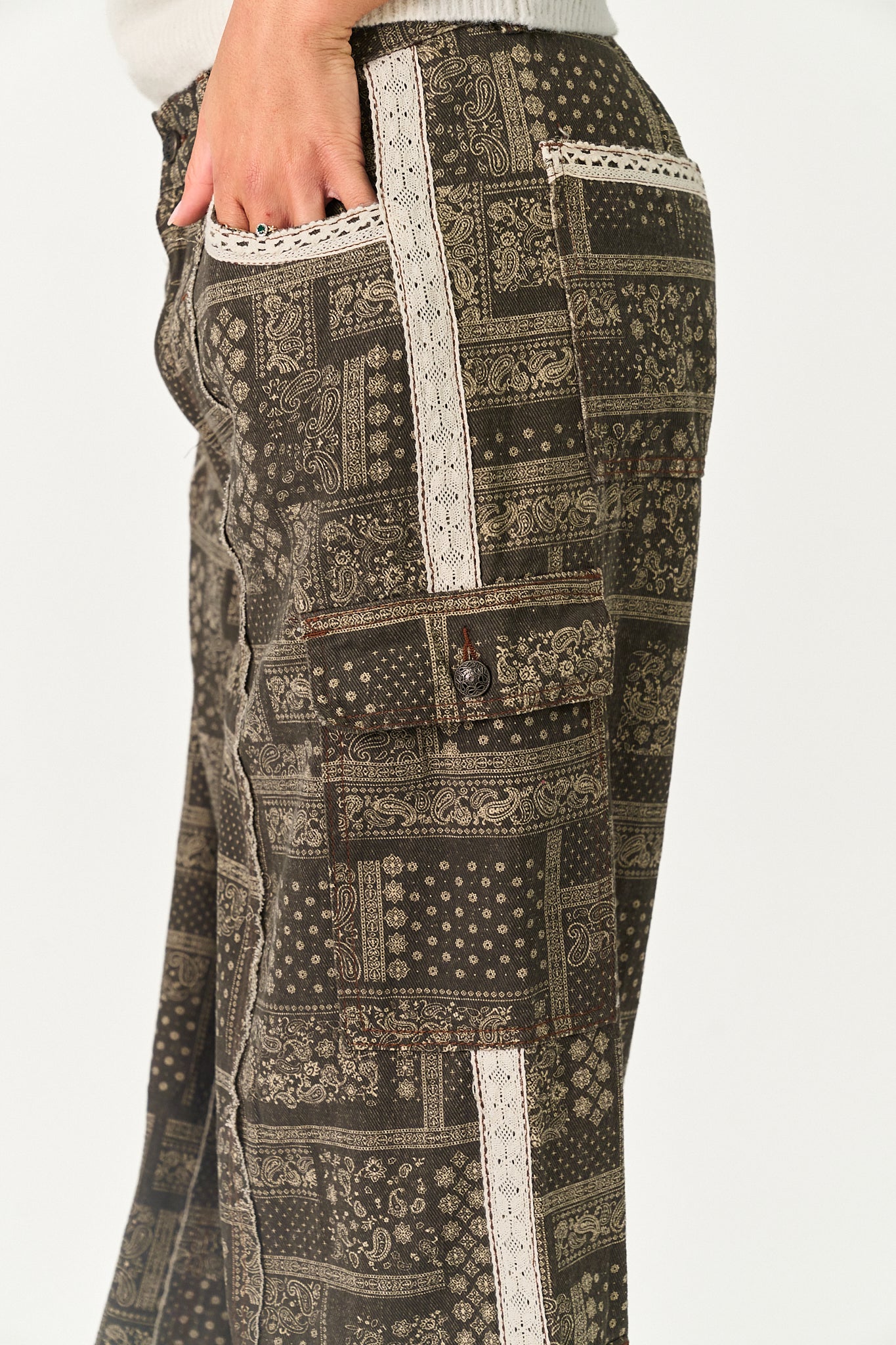 Close-up of a person wearing patterned cargo pants with a dark brown and beige patchwork print and white lace trim, shown against a plain light background.