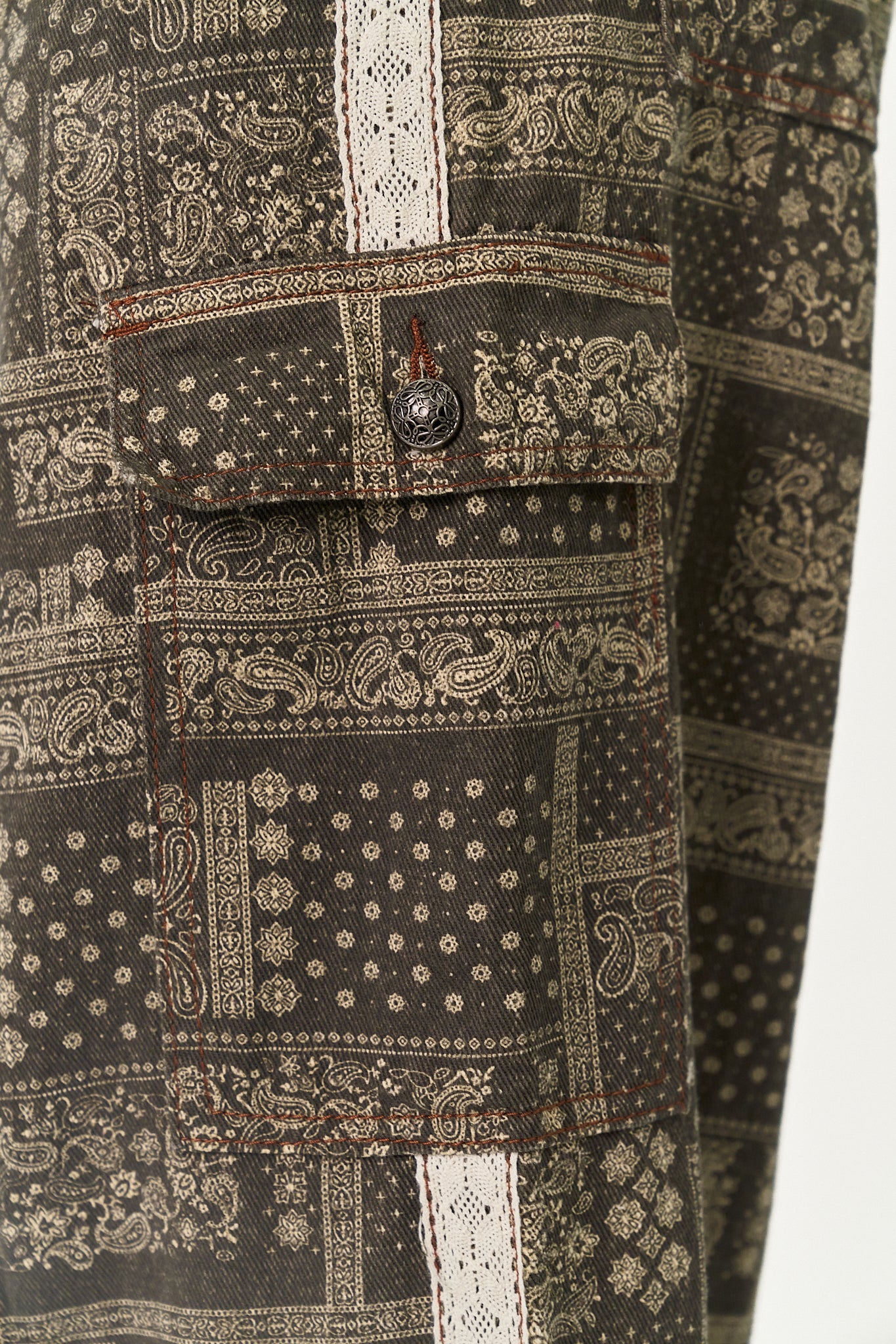 Close-up of a dark brown and beige paisley bandana-patterned fabric with white lace-like vertical stripes and a buttoned flap pocket.