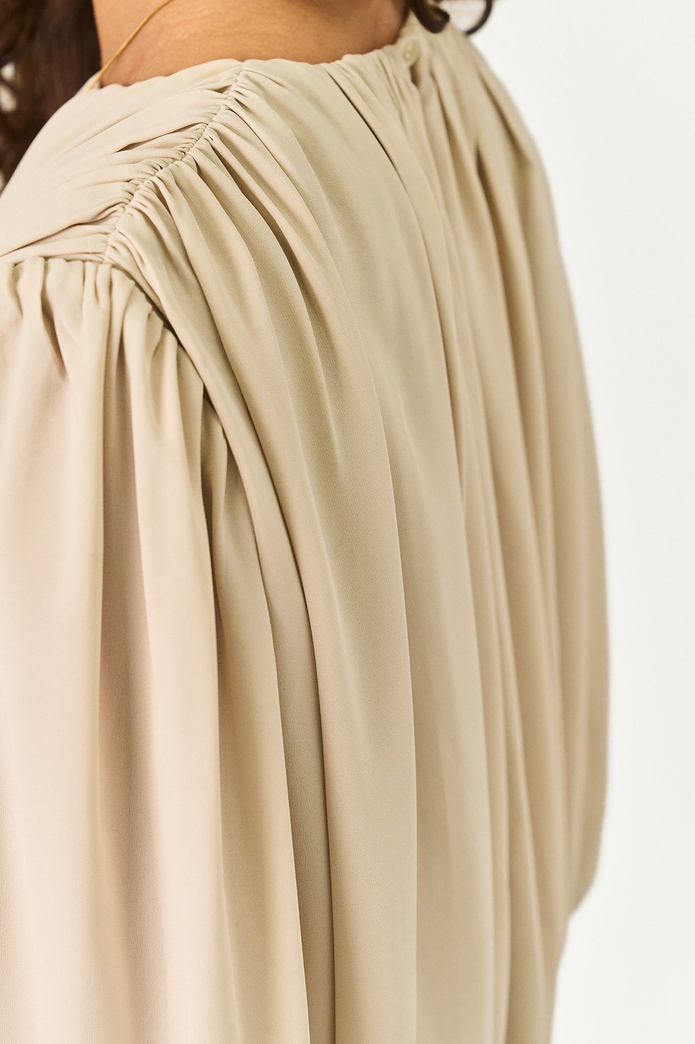 Close-up of a beige or light tan blouse with a gathered, ruched neckline and voluminous long sleeves, shown on a model against a plain white background.