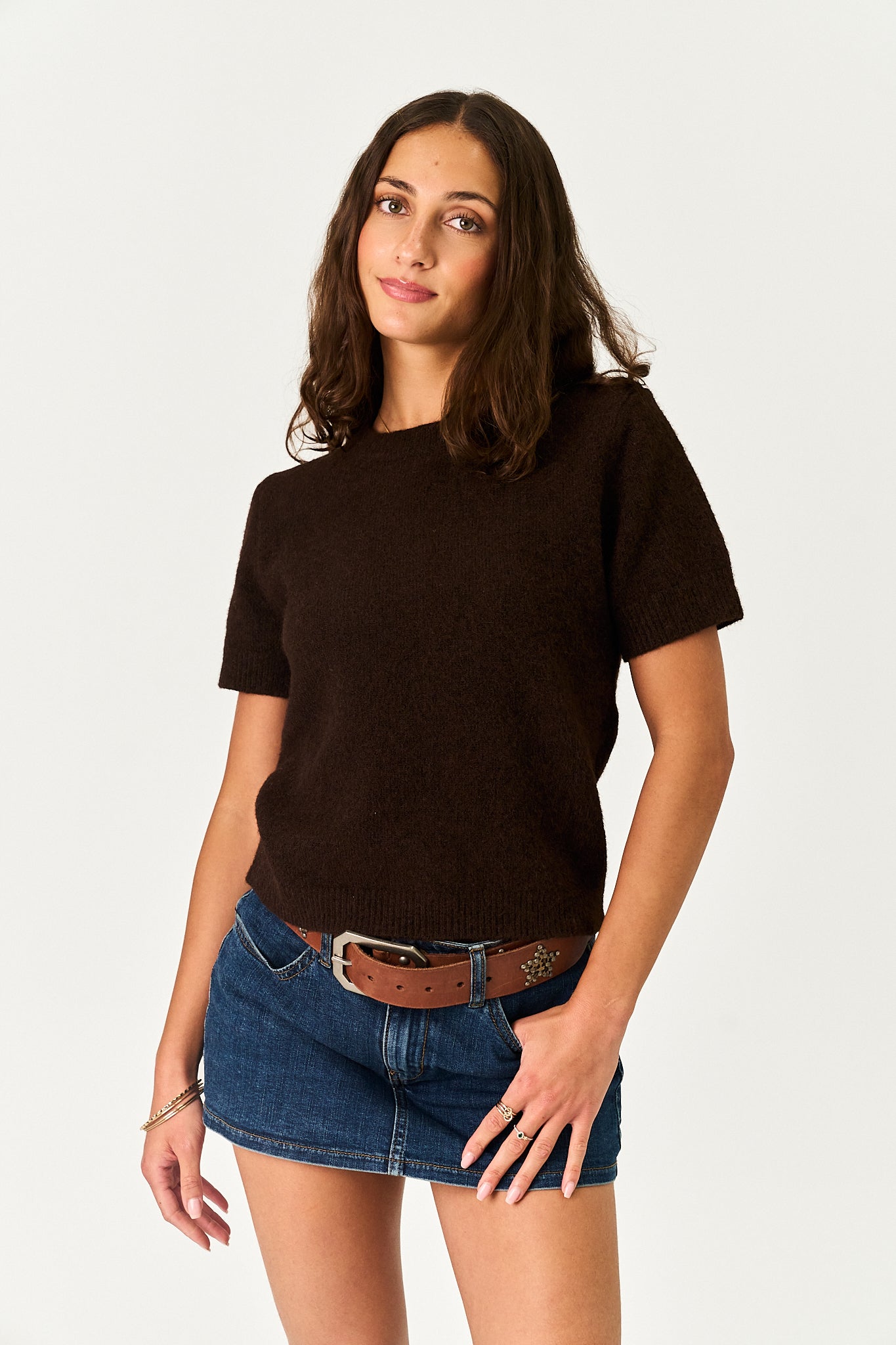 A woman modeling a dark brown short-sleeve knit top with a crew neck, paired with a brown belt and denim mini skirt, against a plain light gray background.