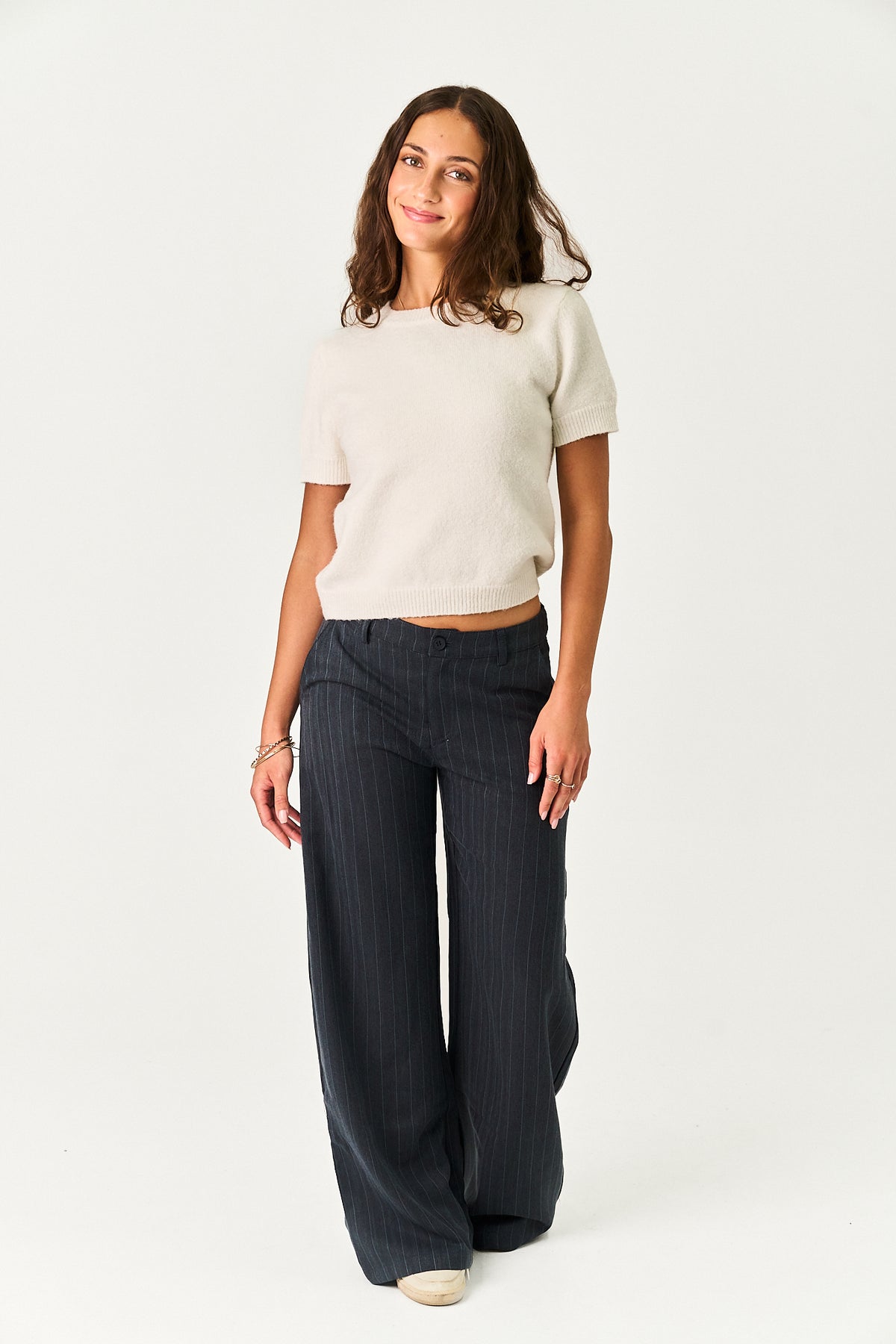 A woman modeling a short-sleeve cream knit crop top and dark navy wide-leg pinstripe trousers against a plain white studio background.