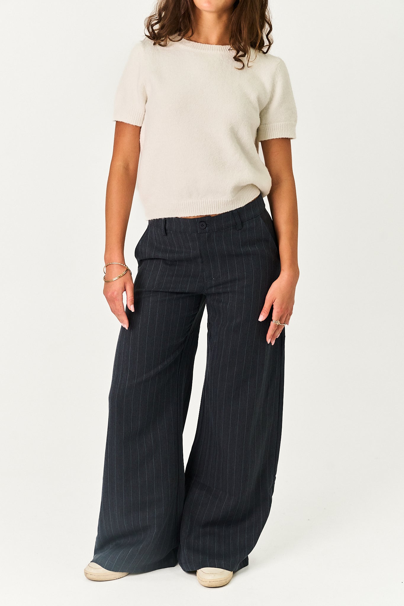 A model wearing a cream short-sleeve knit top and dark navy wide-leg pinstripe trousers, standing against a plain white studio background.