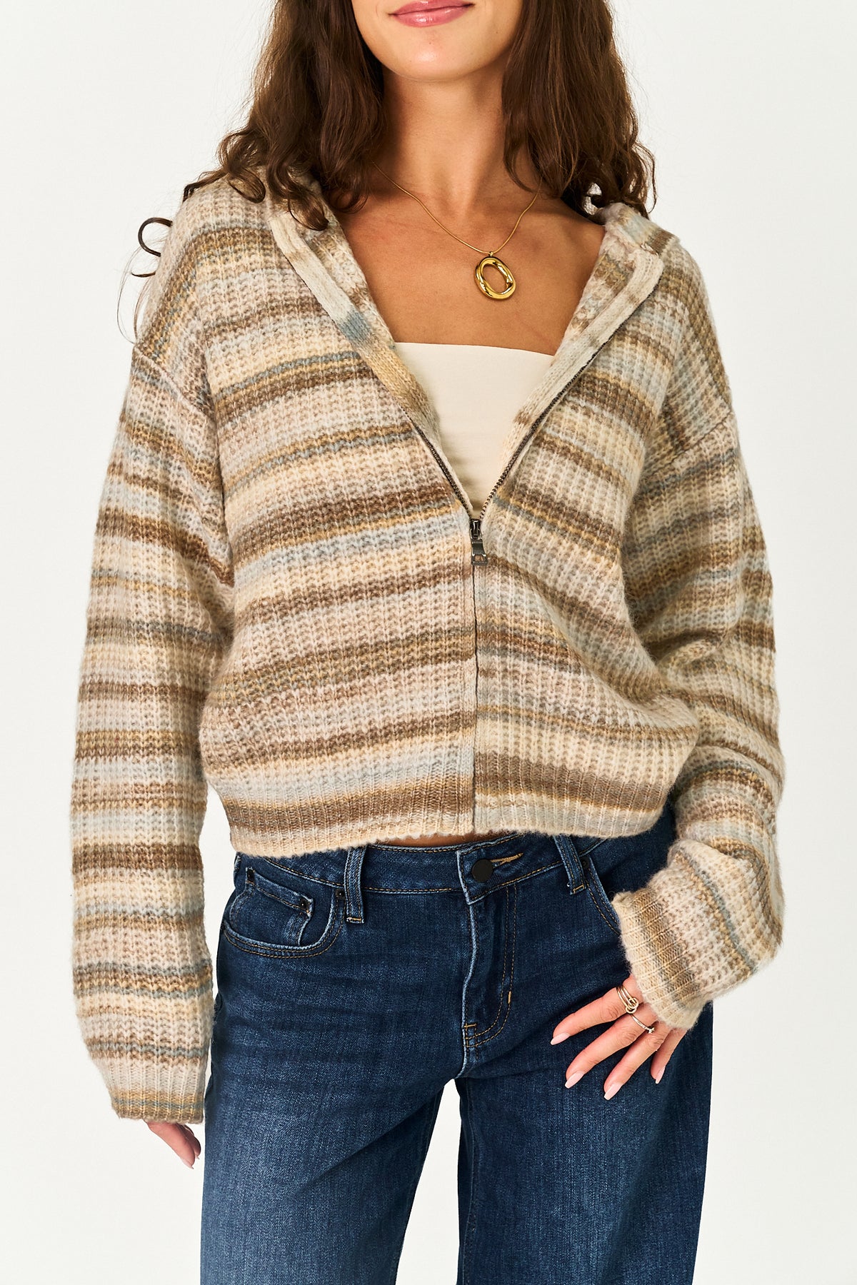 A woman wearing a cropped zip-up hooded sweater with horizontal beige, brown, and tan stripes, styled over a cream top and blue jeans.