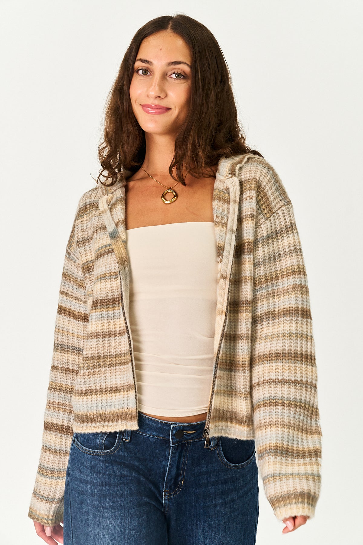 A woman wearing a beige and brown striped knit zip-up hoodie over a cream strapless top and blue jeans, photographed against a plain light gray studio background.
