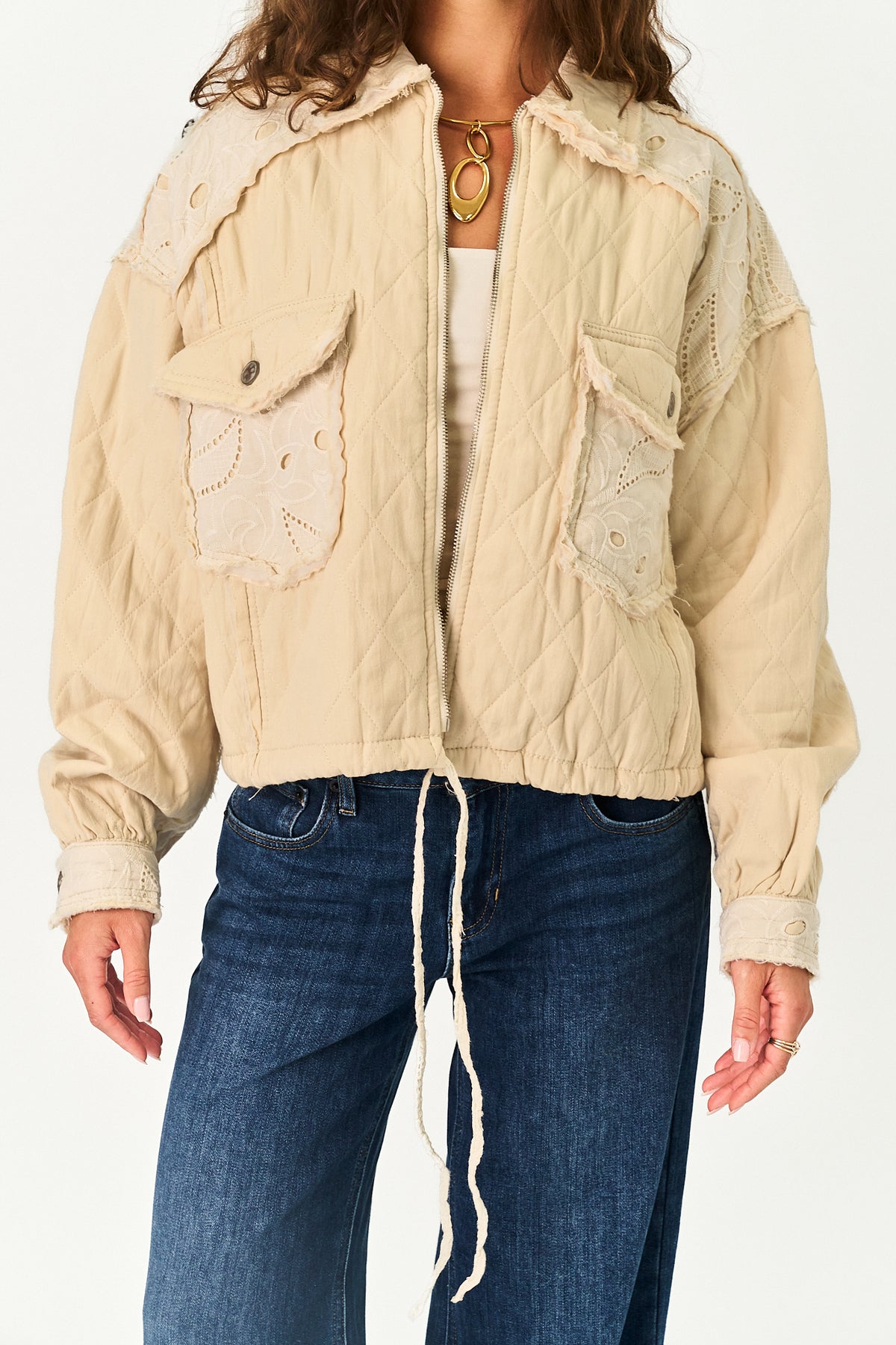 A woman wearing a cream-colored quilted jacket with lace appliques and drawstring details, paired with blue jeans and a gold necklace, against a plain white background.