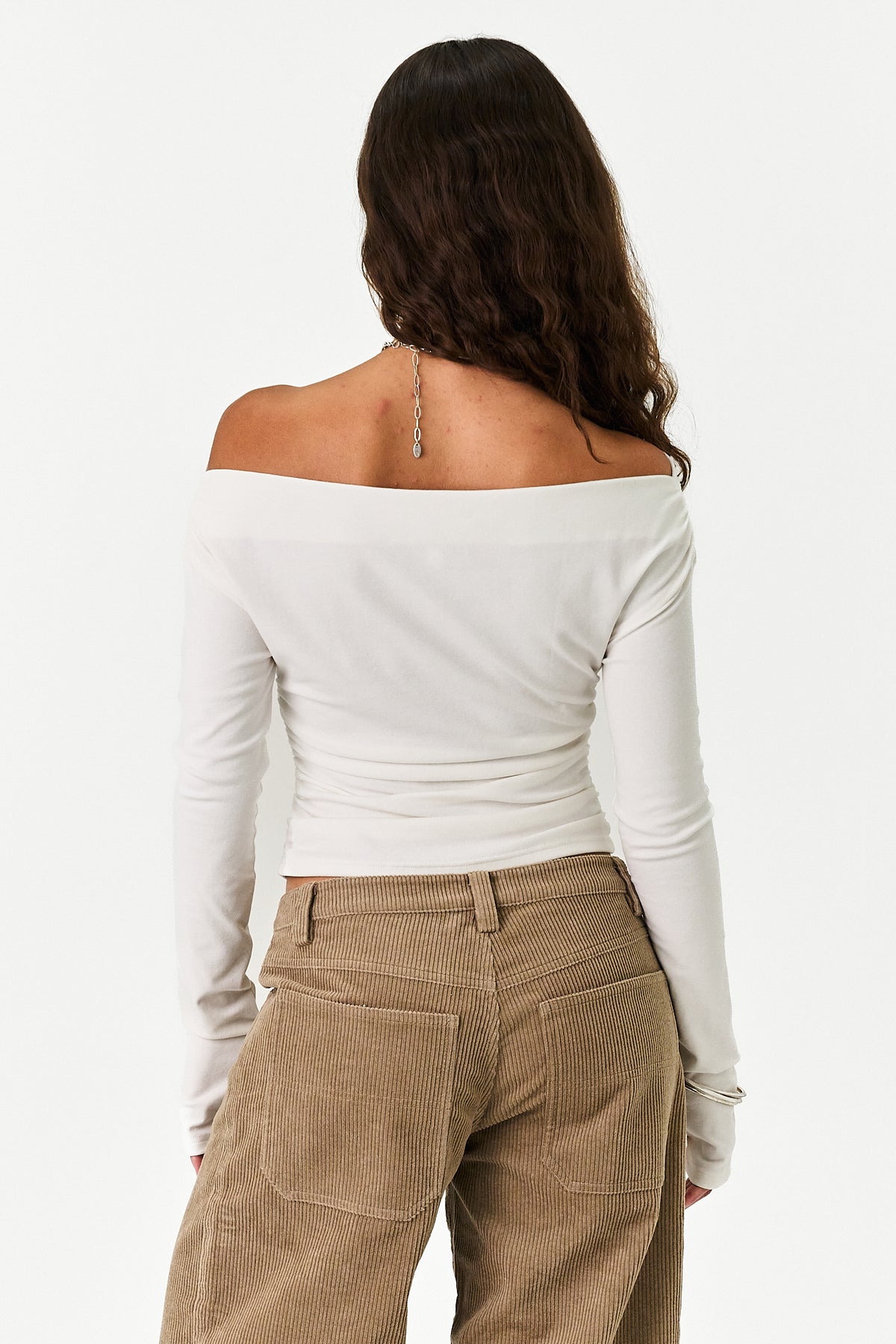 A woman shown from the back wearing a fitted off-the-shoulder long-sleeve top in off-white and tan corduroy pants, against a plain white studio background.