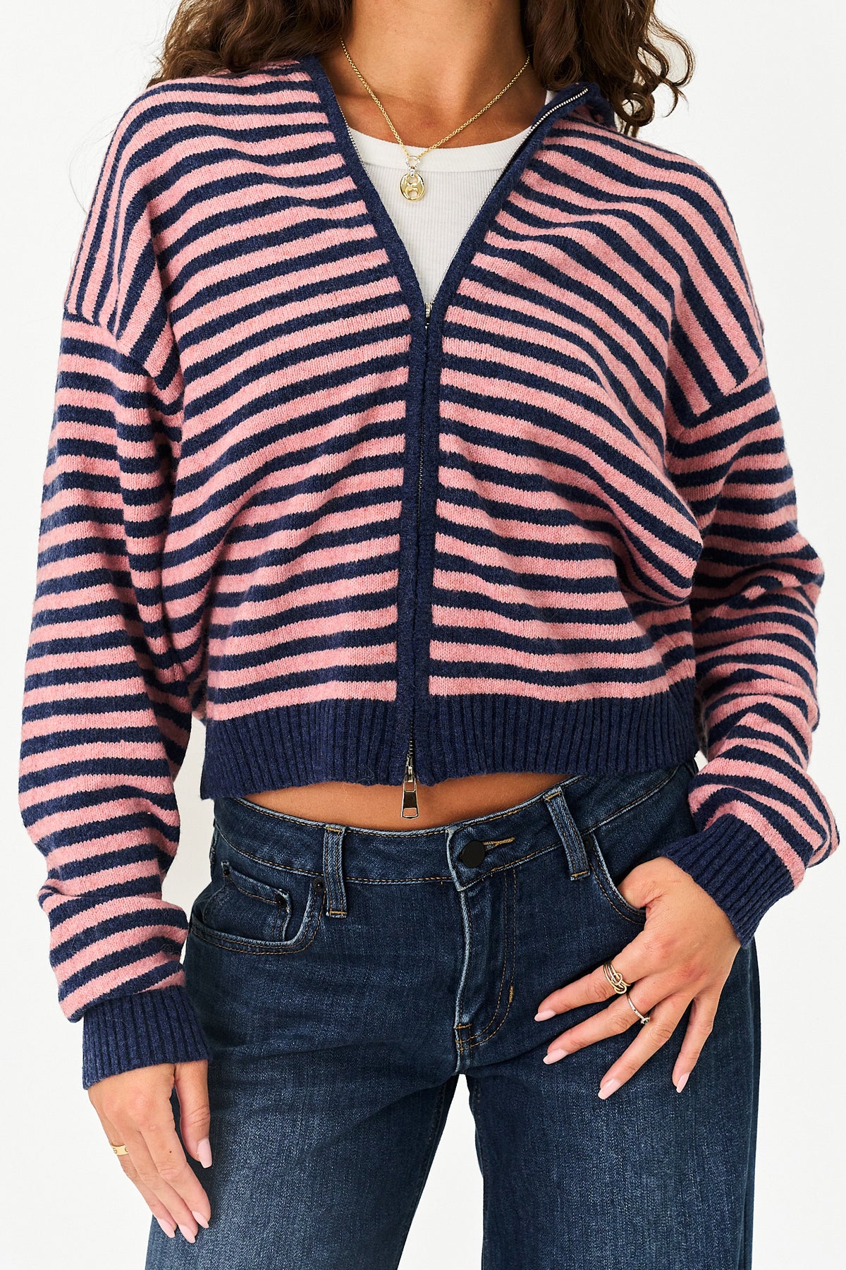 A woman wearing a cropped zip-up cardigan with horizontal pink and navy stripes, paired with dark blue jeans and a white undershirt.