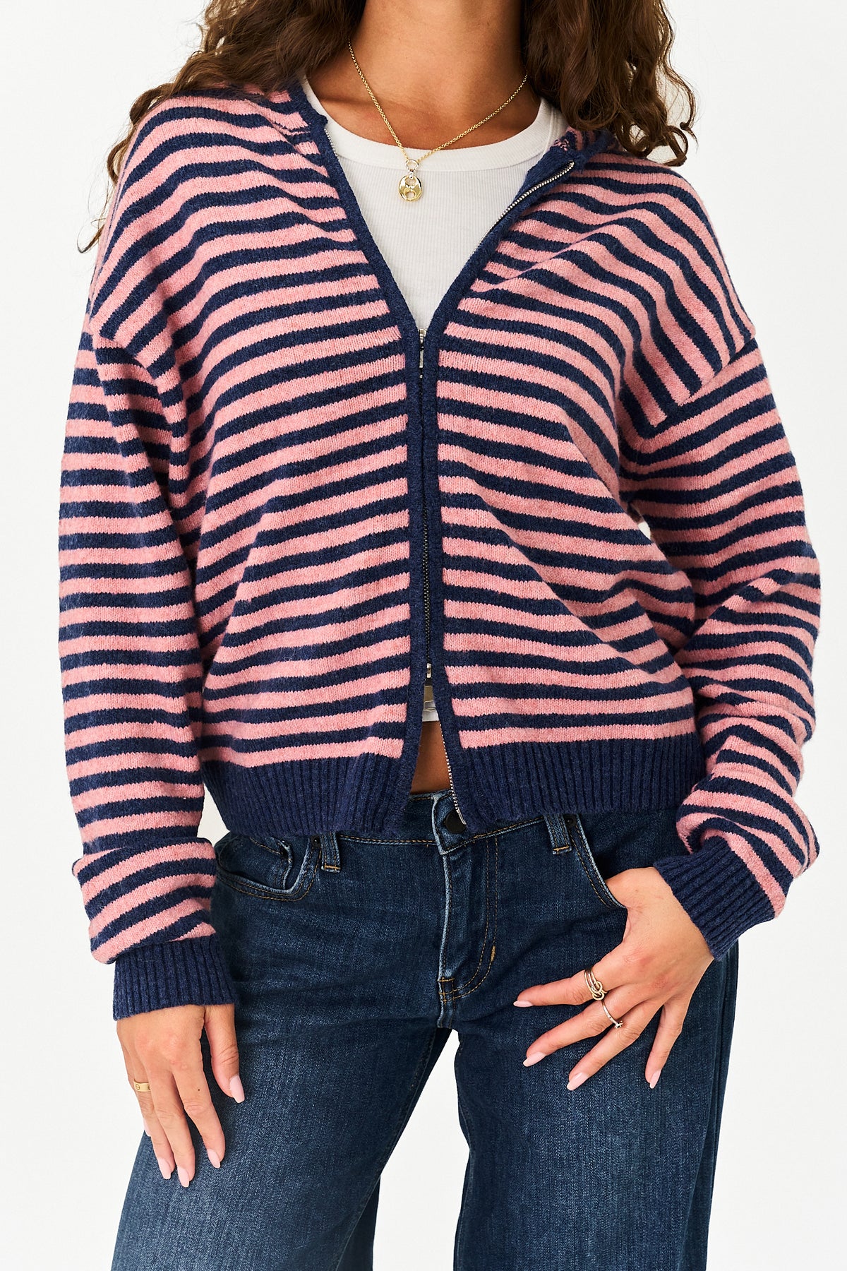A woman wearing a pink and navy blue horizontally striped zip-up cardigan over a white top and dark blue jeans, shown from the shoulders to the upper thighs against a plain light background.