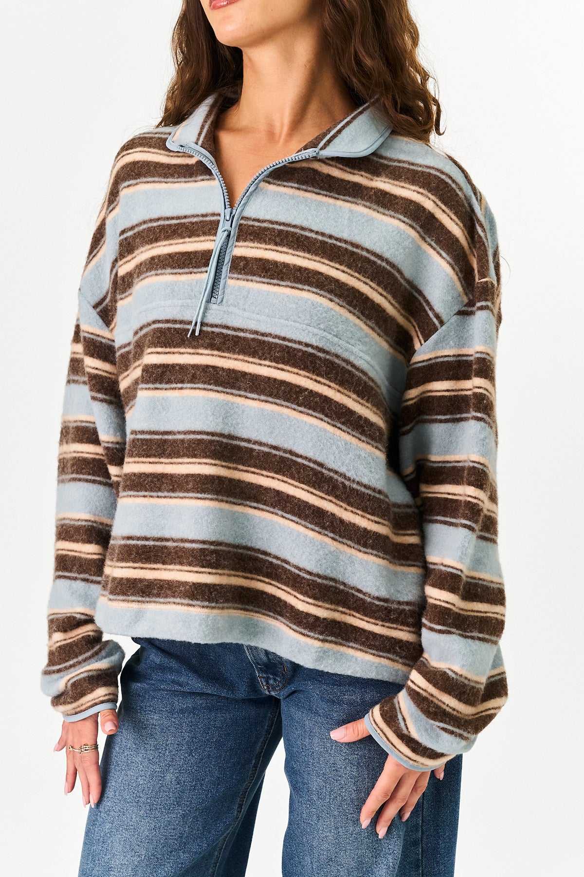 A woman wearing a long-sleeve quarter-zip pullover sweater with horizontal stripes in light blue, brown, and beige, paired with blue jeans against a plain white background.