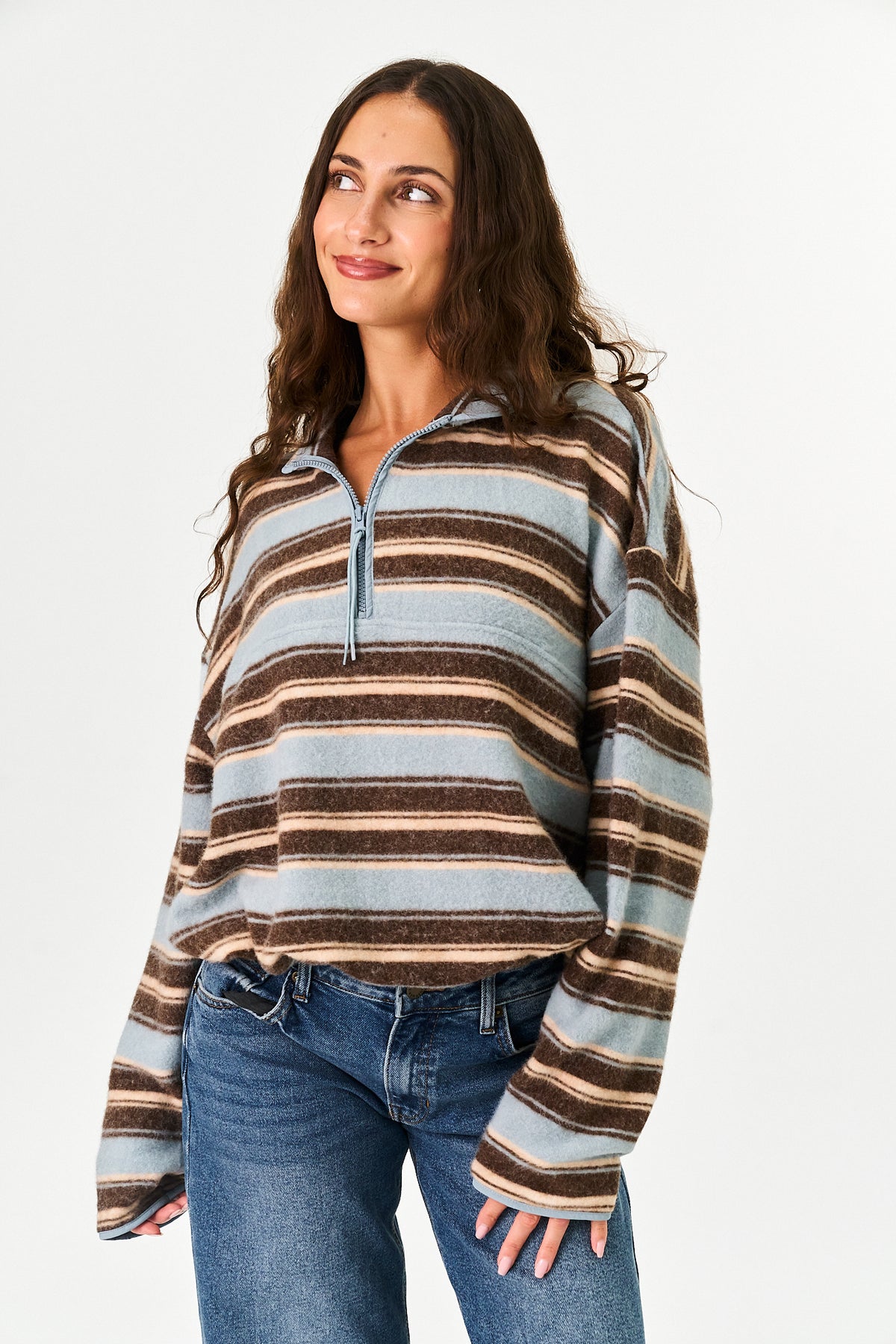A woman wearing a long-sleeve quarter-zip knit sweater with horizontal stripes in brown, beige, and light blue, paired with blue jeans, against a plain white studio background.