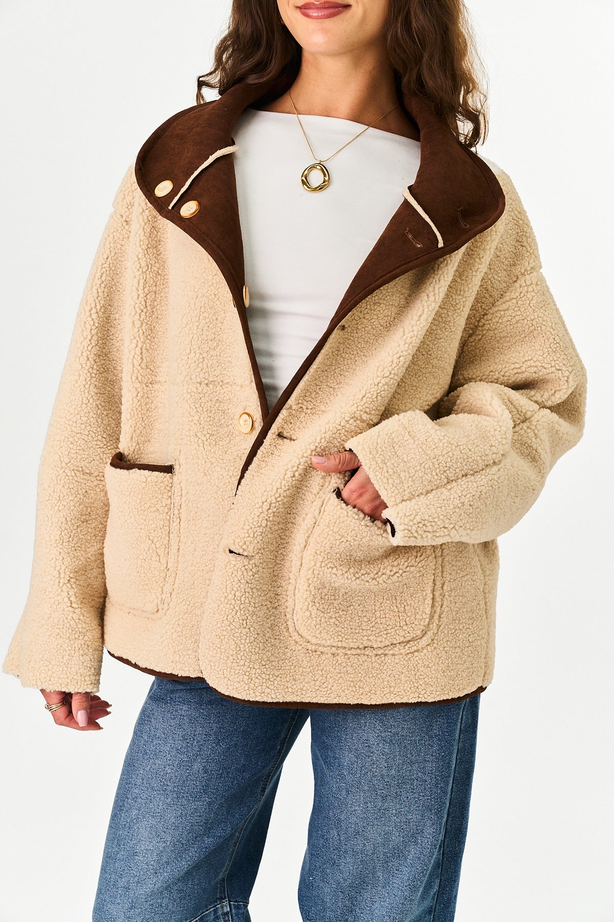 A person wearing a light brown sherpa-like jacket with a brown suede collar and contrast beige piping, paired with blue jeans, against a plain white studio background.